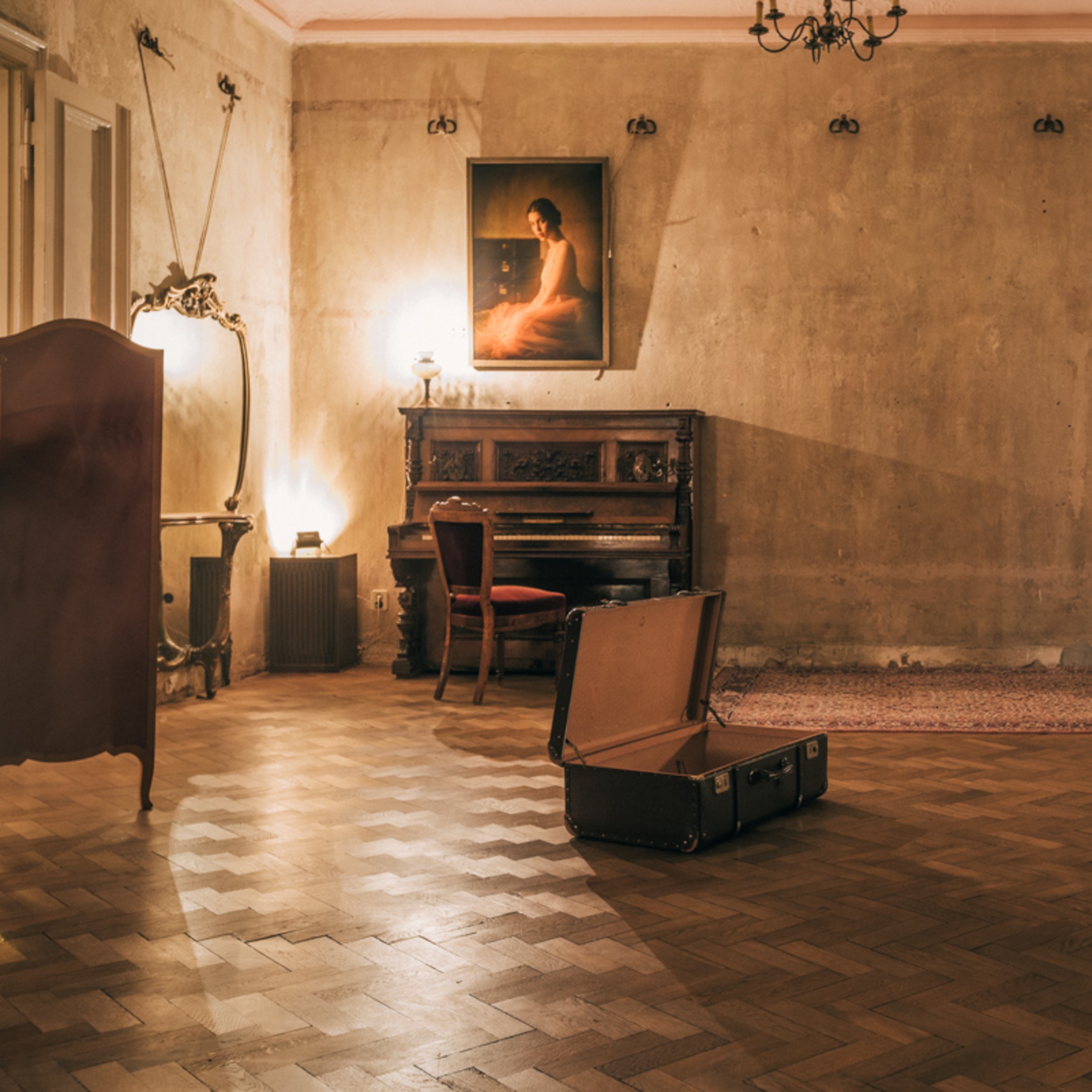 A vintage suitcase lies open on the wooden floor of a dimly lit, empty room featuring a piano, a patterned armchair, and a portrait hanging on the wall.