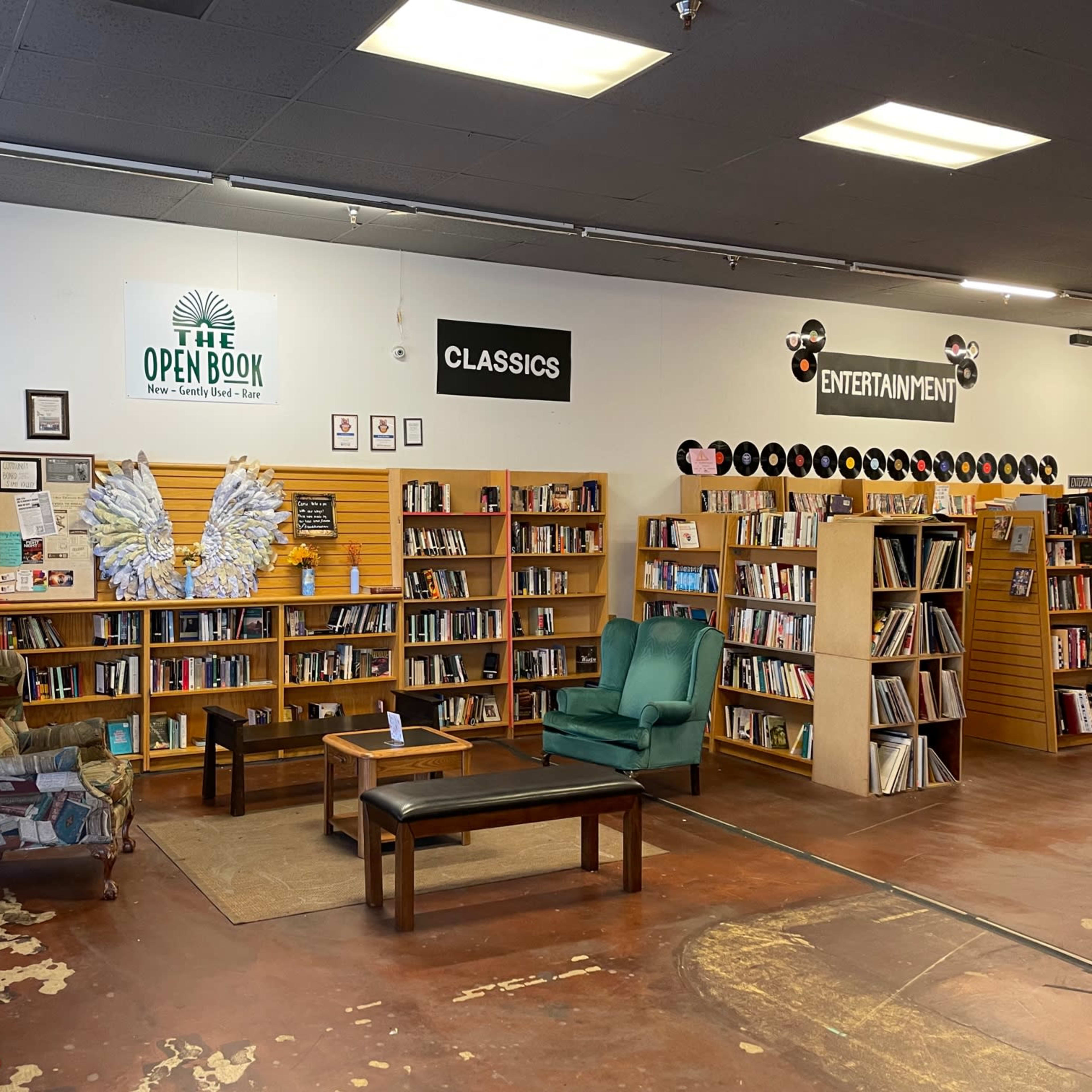A cozy library space with wooden bookshelves filled with books, a seating area with two chairs and a small table, and signs indicating sections like "The Open Book," "Classics," and "Entertainment."