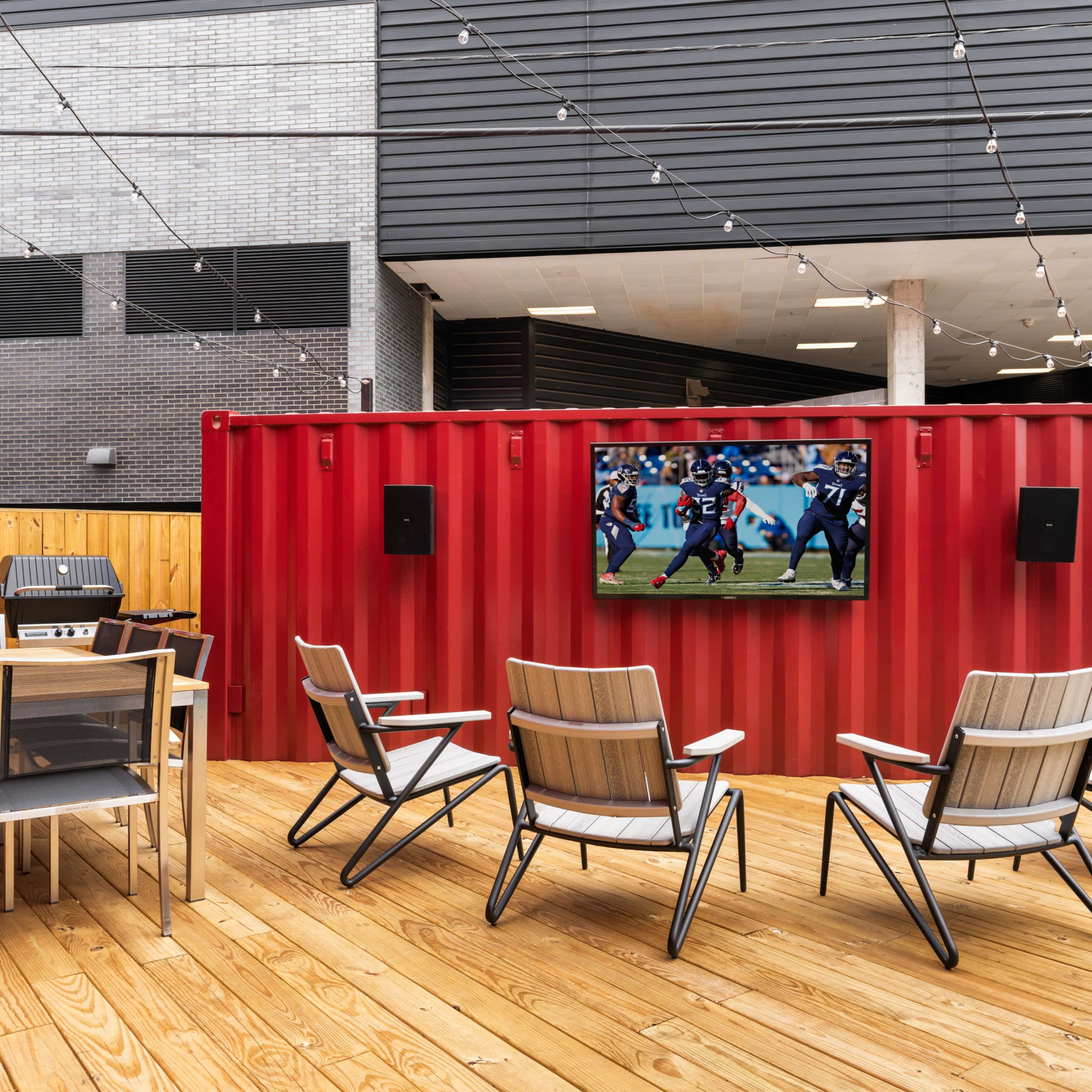 A spacious outdoor area features a red shipping container wall with a mounted television, surrounded by seating and a dining table on wooden decking.