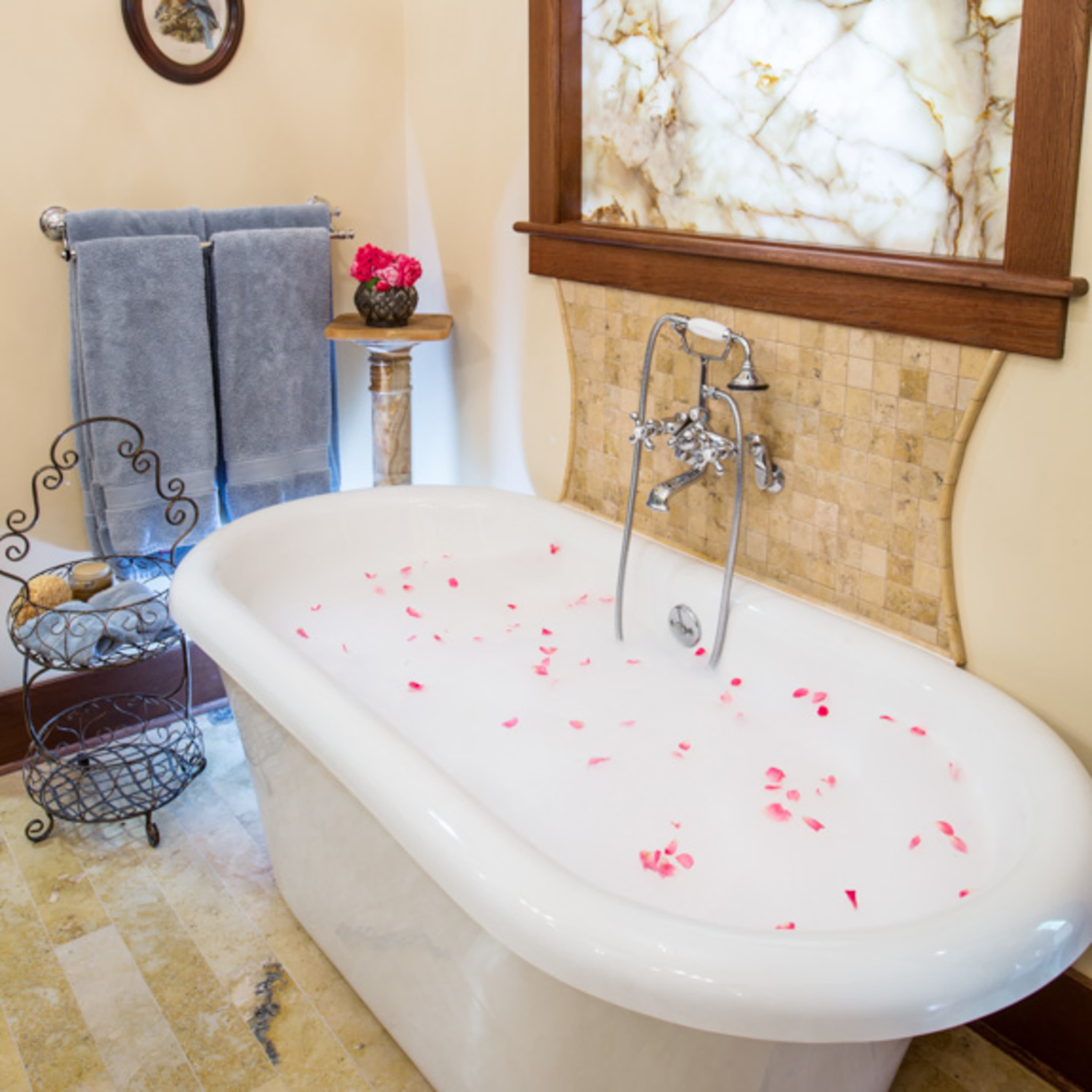 A bathtub filled with water and rose petals, surrounded by a stone and marble wall, a decorative wall-mounted mirror, and a towel rack with a gray towel.
