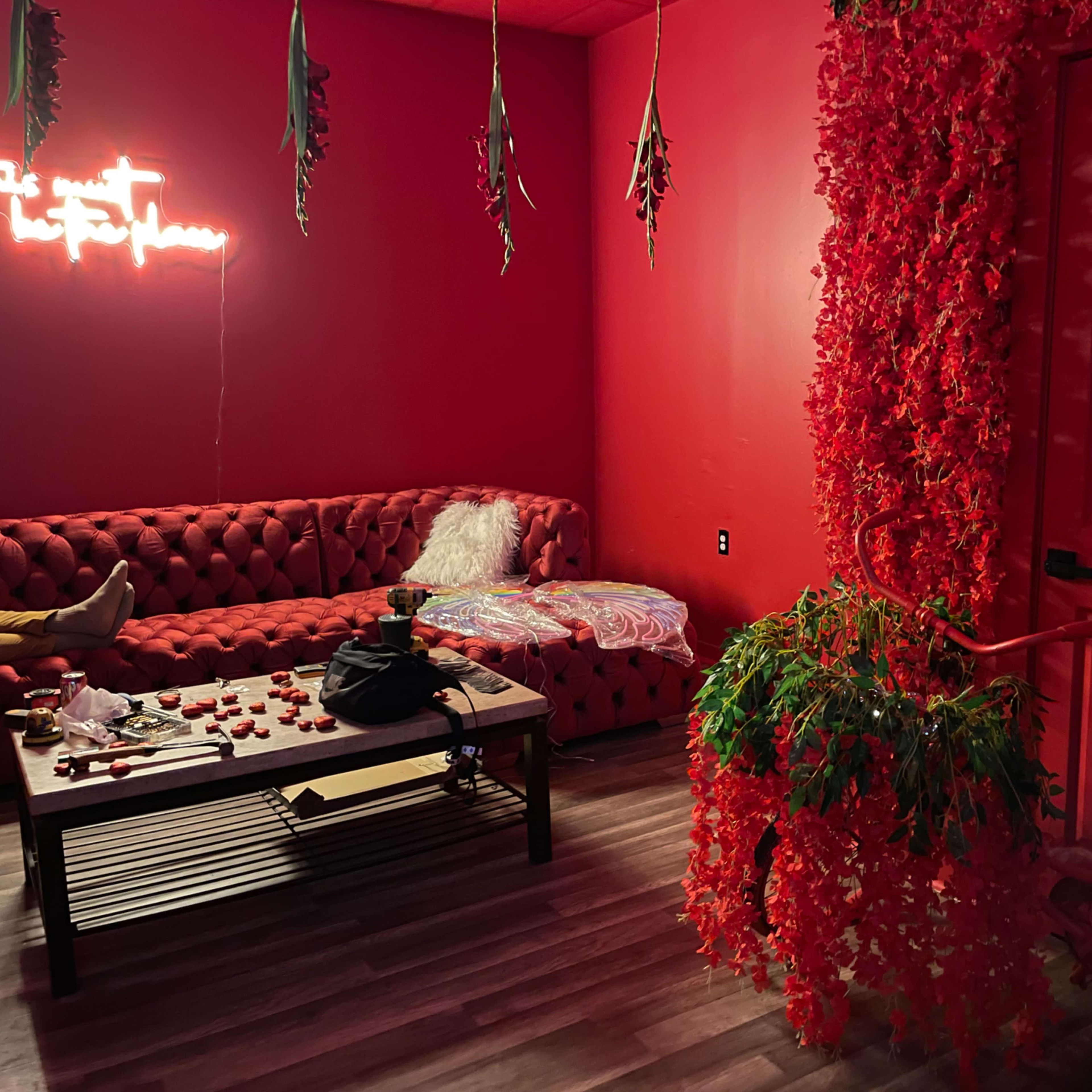 The room features red walls, a red tufted couch, a coffee table scattered with rose petals, and a bicycle adorned with artificial flowers at the entrance.