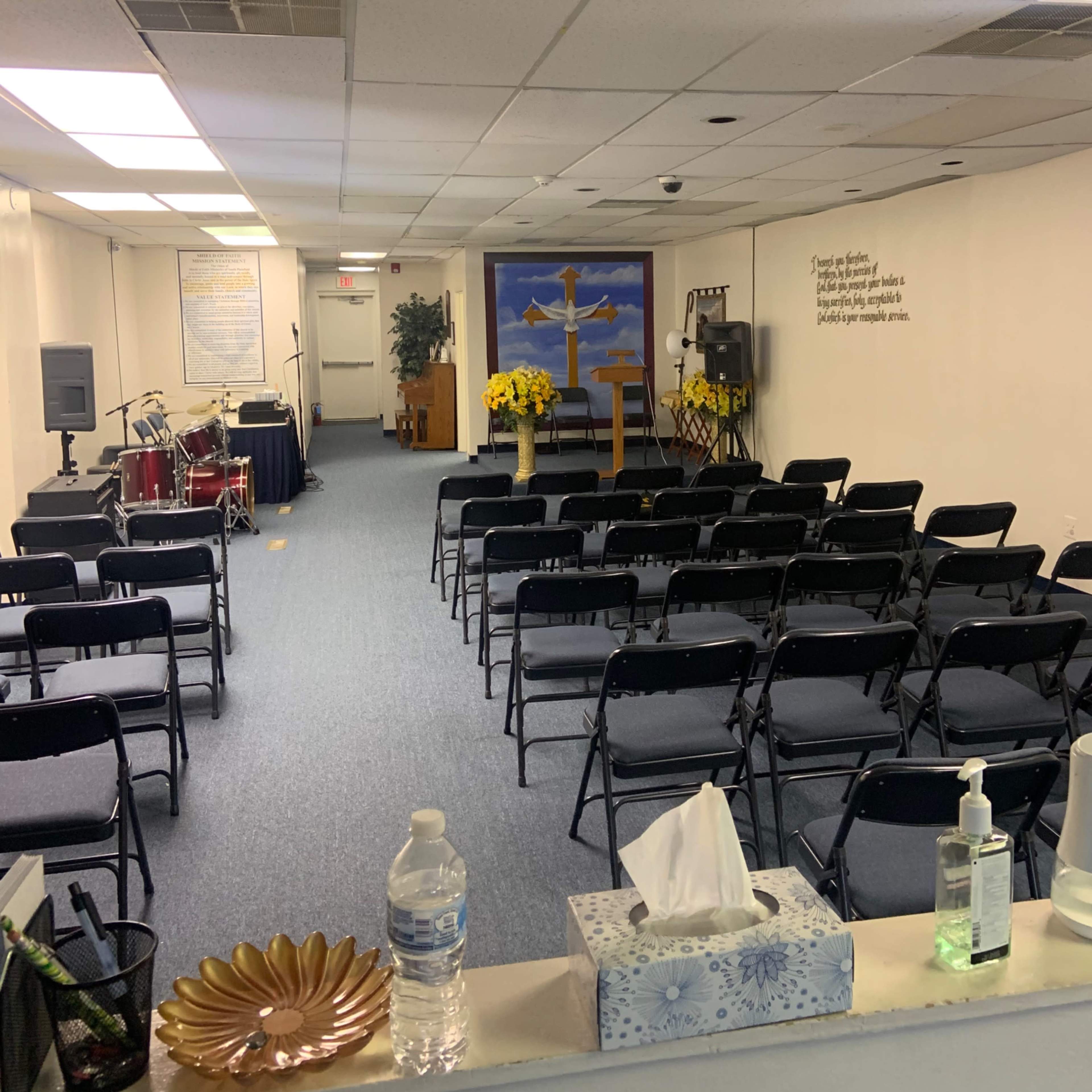A modest indoor space set up for a gathering, featuring rows of black chairs facing a stage with a microphone, drums, and a backdrop displaying a cross and a scenic painting.