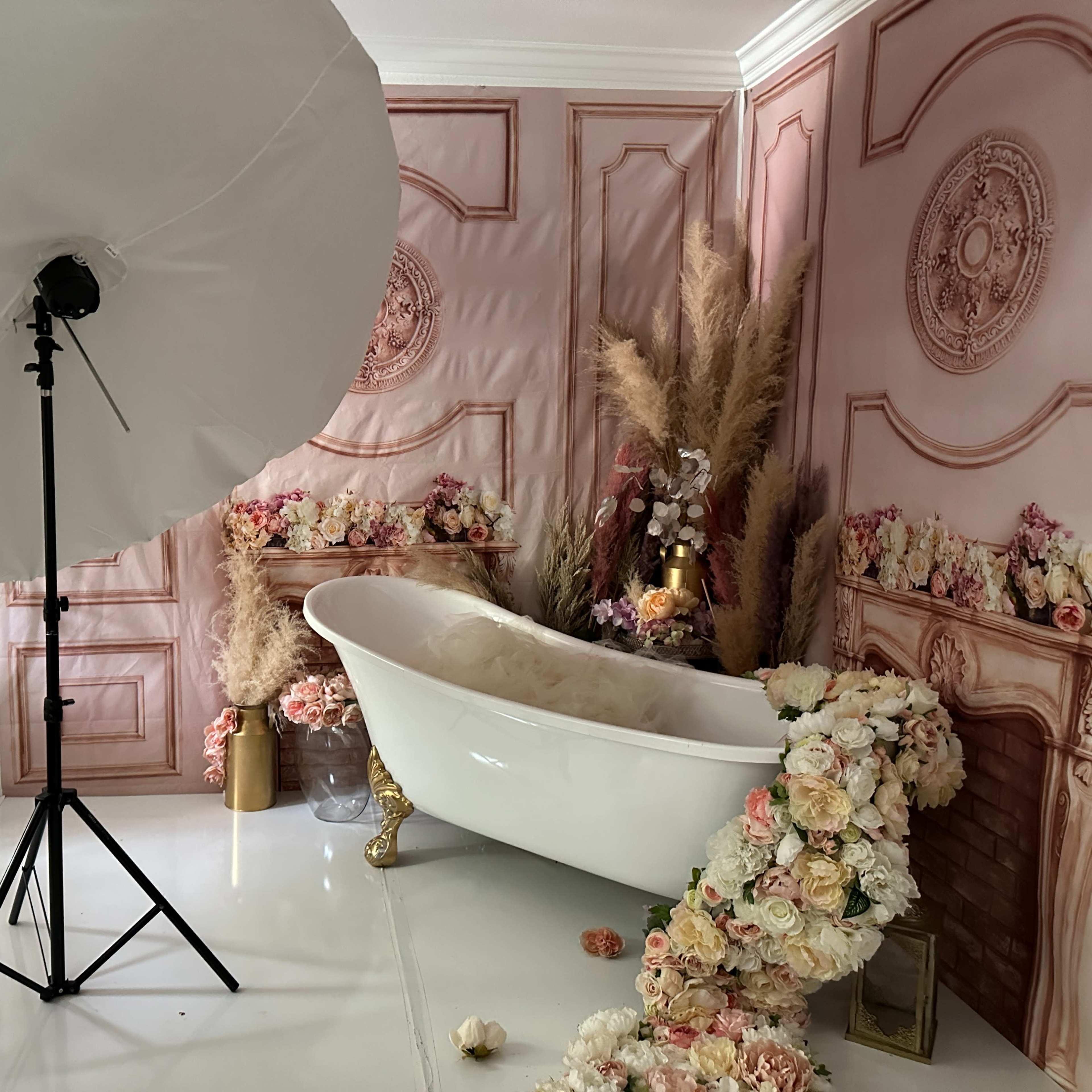 A decorated bathroom featuring a white clawfoot bathtub surrounded by floral arrangements and a large photography umbrella.