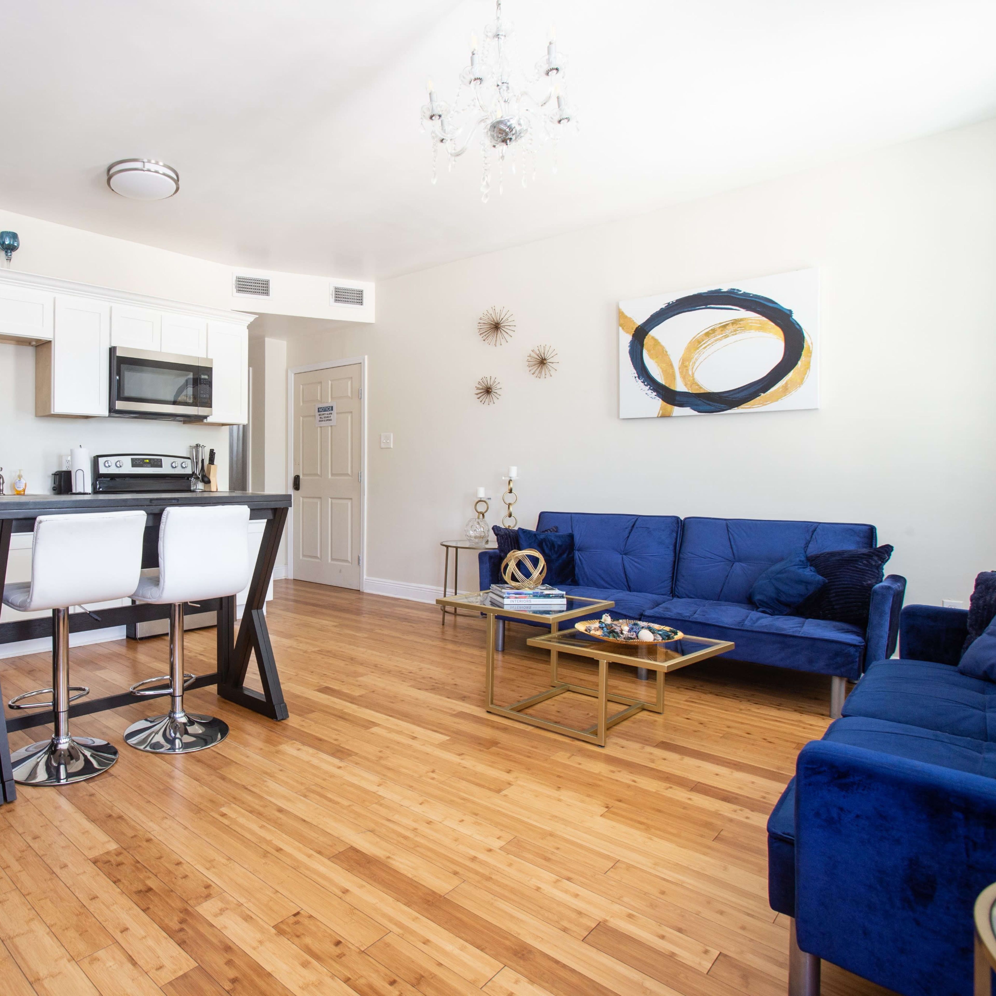 A modern living space featuring a kitchen bar with white stools, a dark refrigerator, and a seating area with blue upholstery and a glass coffee table.