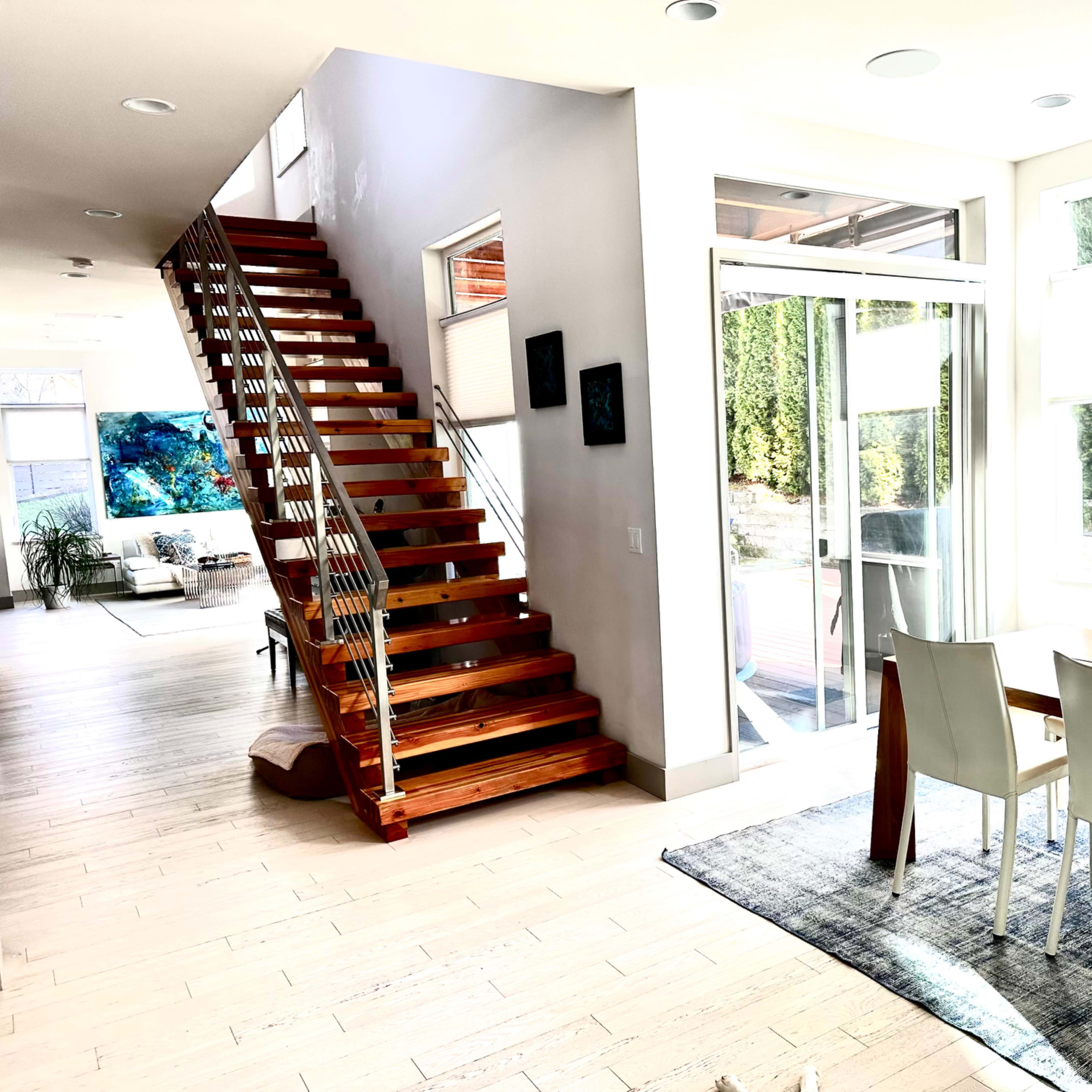 A modern interior features a wooden staircase leading to an upper level, with a dining area and large windows offering natural light.