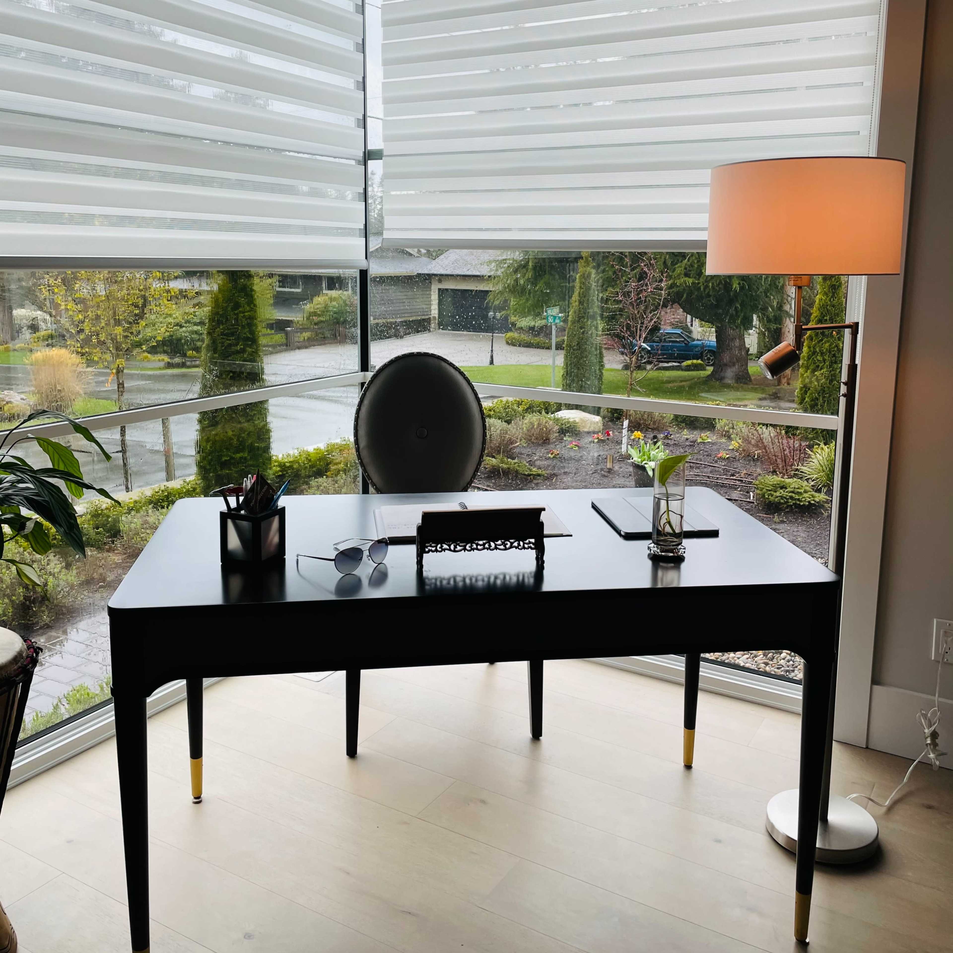 A black desk with a chair is positioned in front of large windows featuring sheer blinds, overlooking a landscaped garden and driveway.