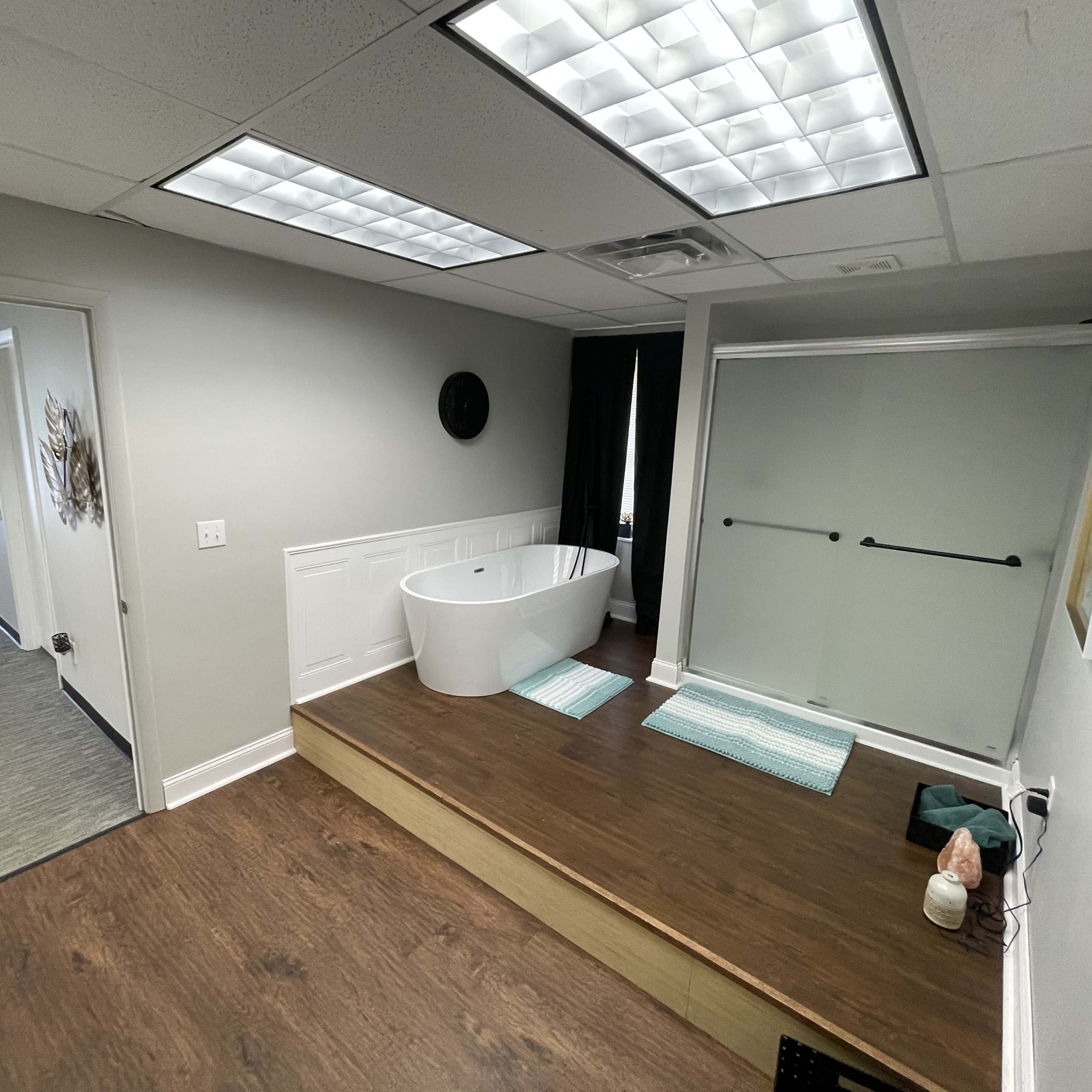A modern bathroom with a freestanding bathtub on a wooden platform, a glass shower enclosure, and two bath mats.