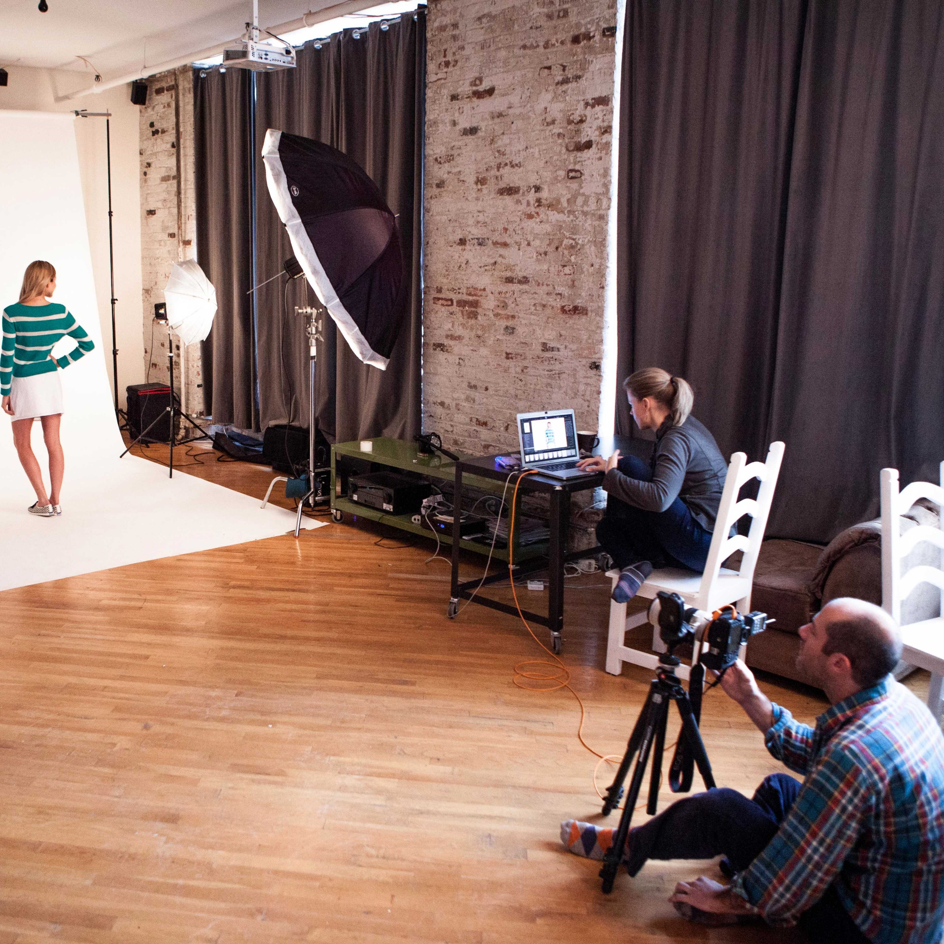 A model poses on a white backdrop in a photography studio while a photographer works at a computer nearby.