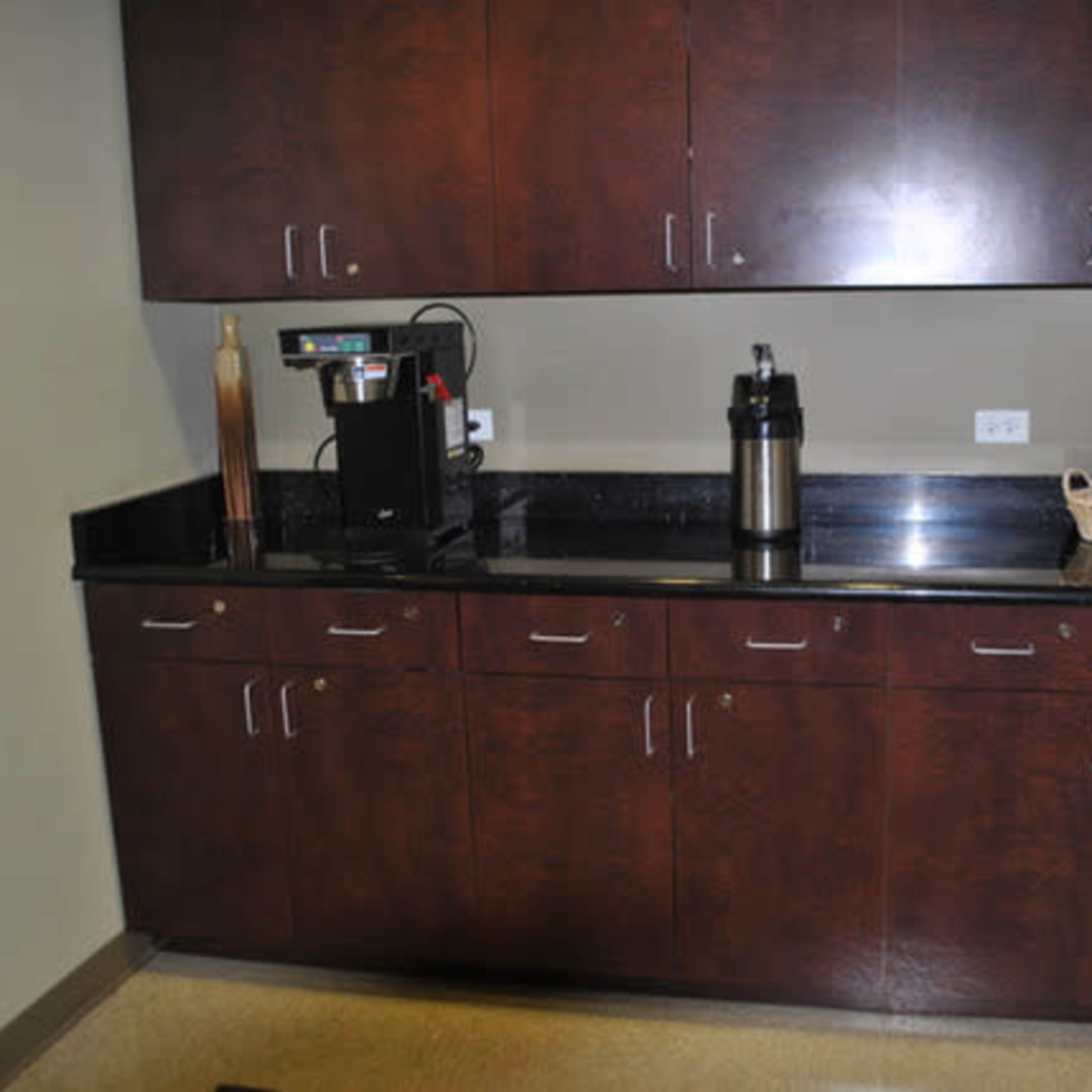 A kitchen area with dark wood cabinetry above a black countertop, featuring a coffee maker and a thermos.