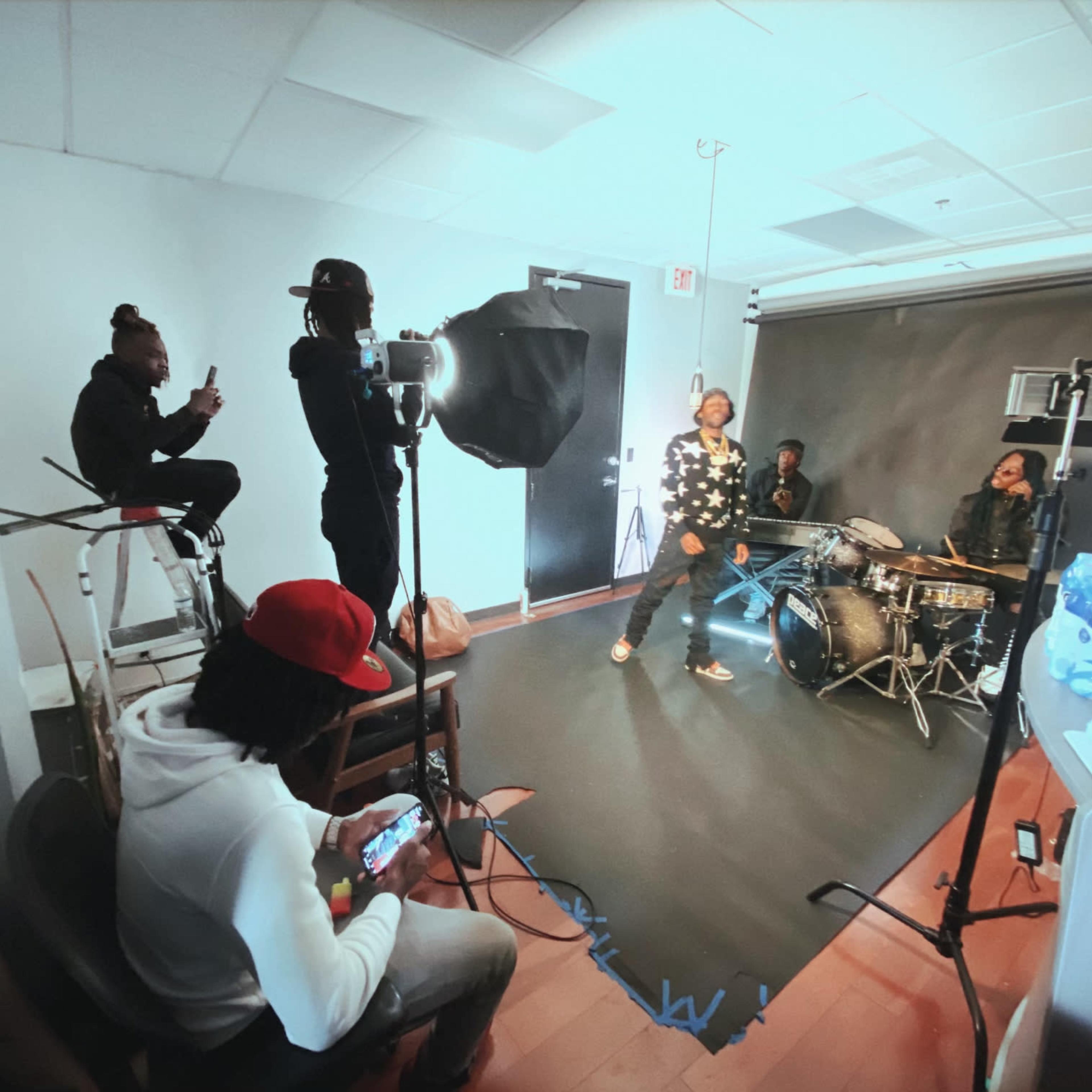 A group of individuals is engaged in a music recording session in a studio, with one person performing on drums and others observing and recording.