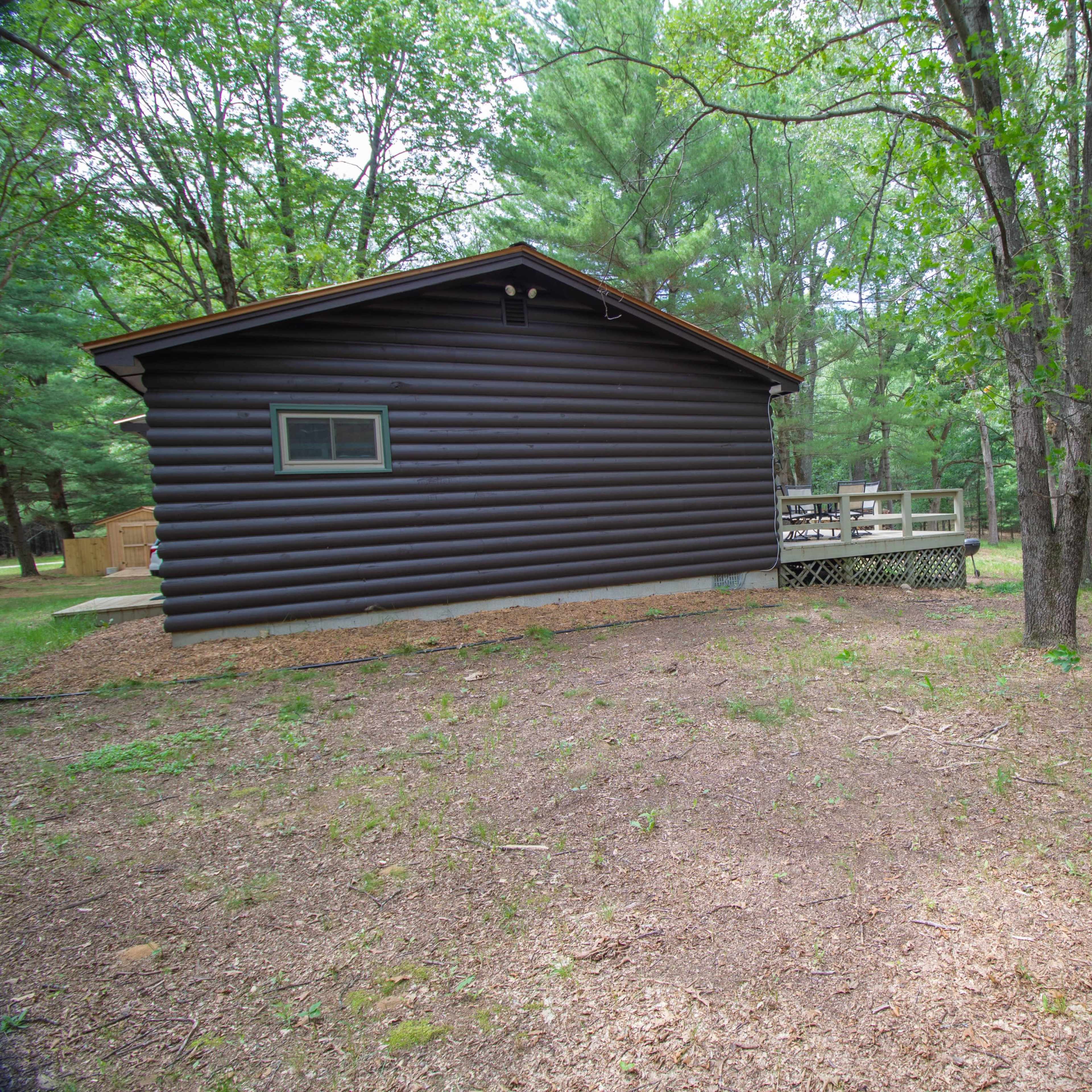 A log cabin is situated among trees in a wooded area, featuring a deck at the rear.