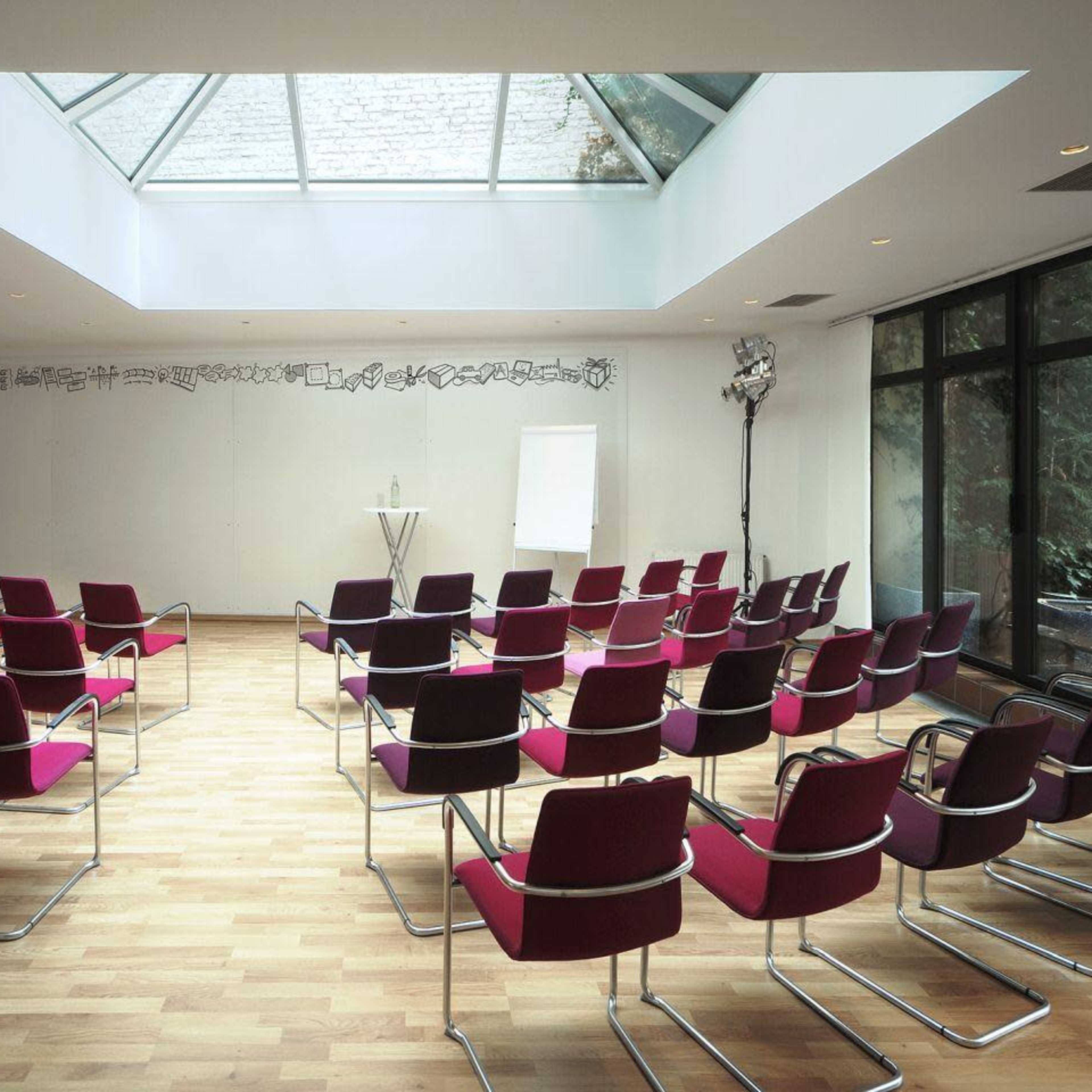 A modern conference room arranged with red and black chairs facing a whiteboard under a skylight.