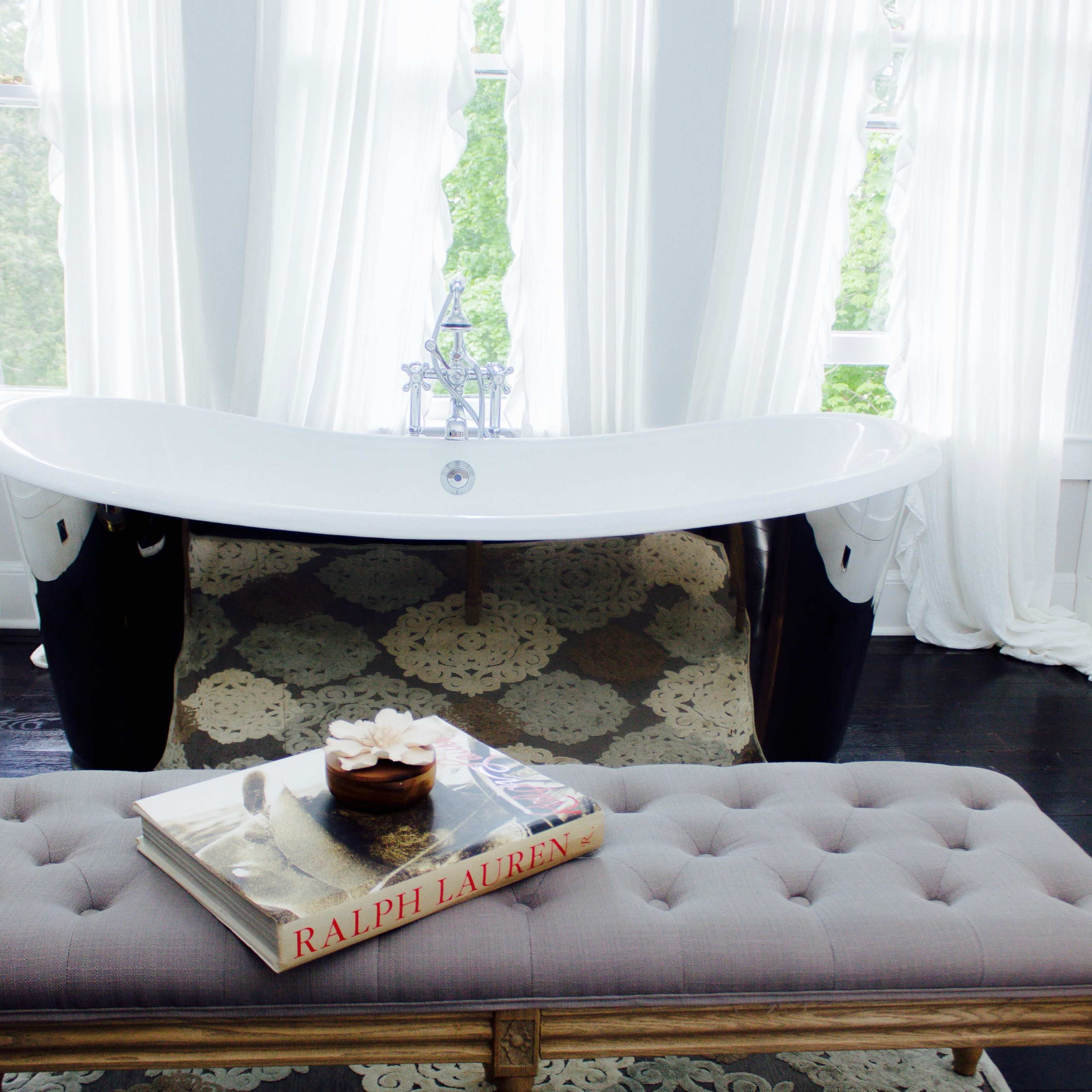 A modern black and white freestanding bathtub is positioned in a bright, airy bathroom with large windows draped in white curtains and a tufted bench holding a styled magazine.