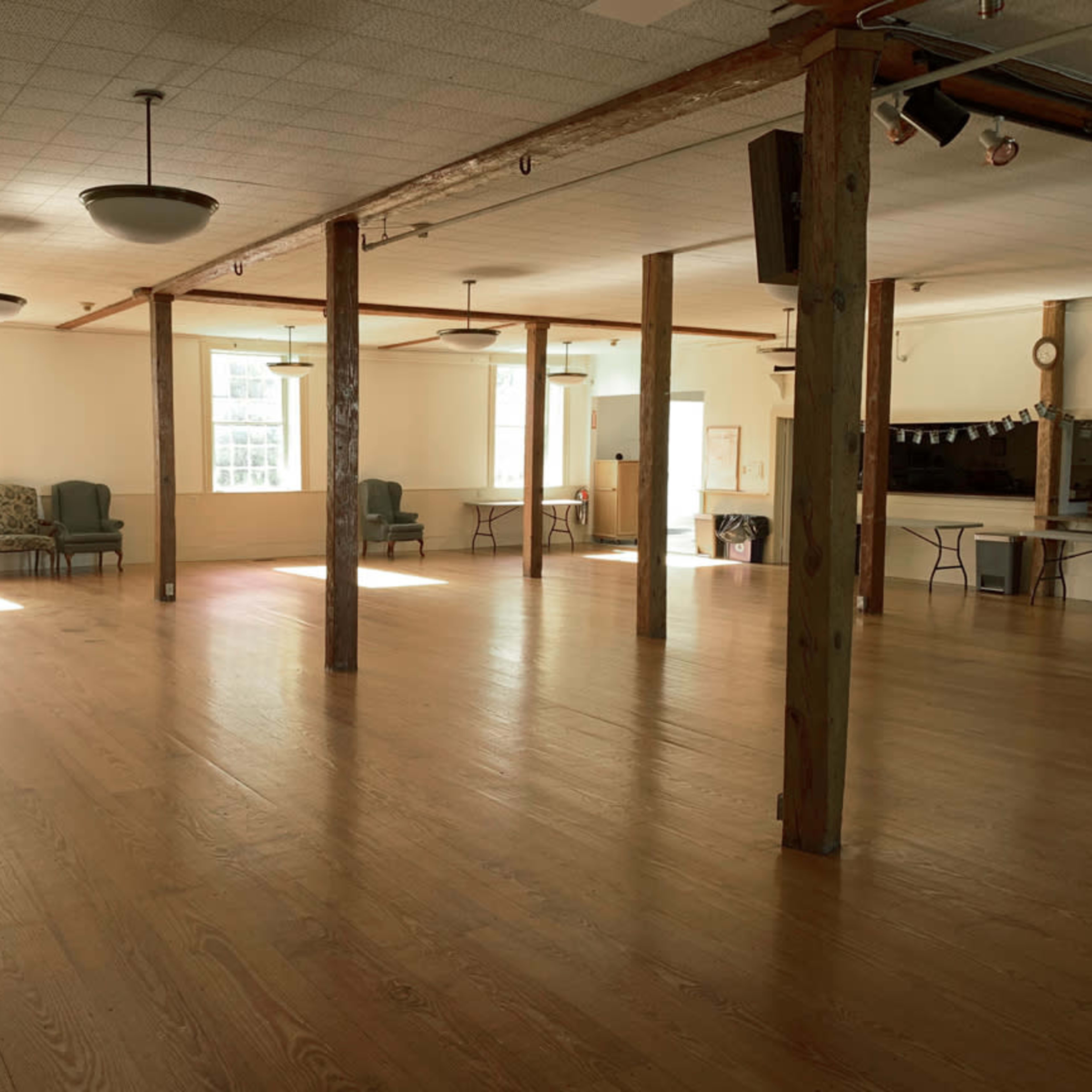 A spacious, well-lit community hall with wooden flooring and several tables and chairs arranged throughout.