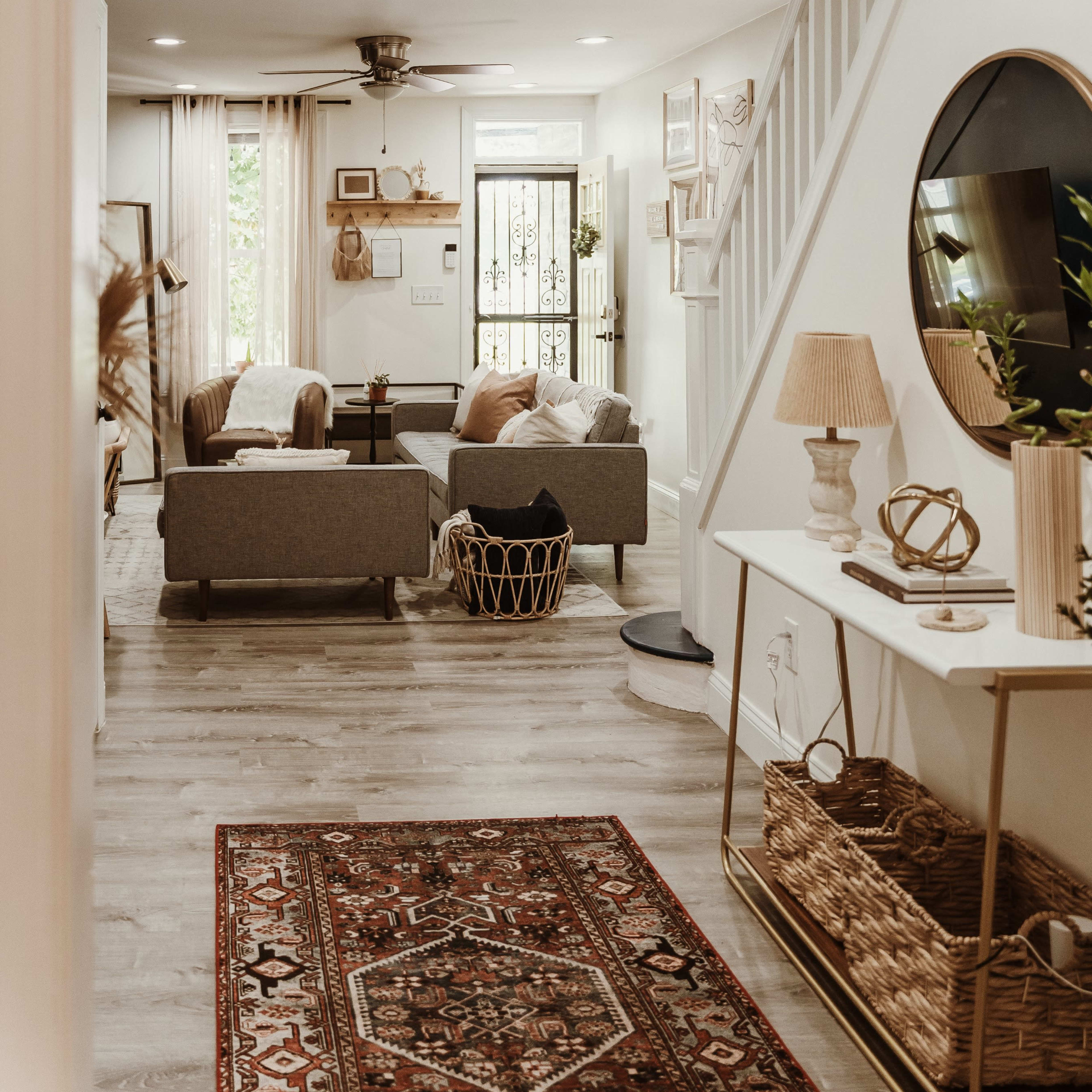A well-lit living room featuring gray sofas, a decorative rug, and a staircase, with a view from a hallway.