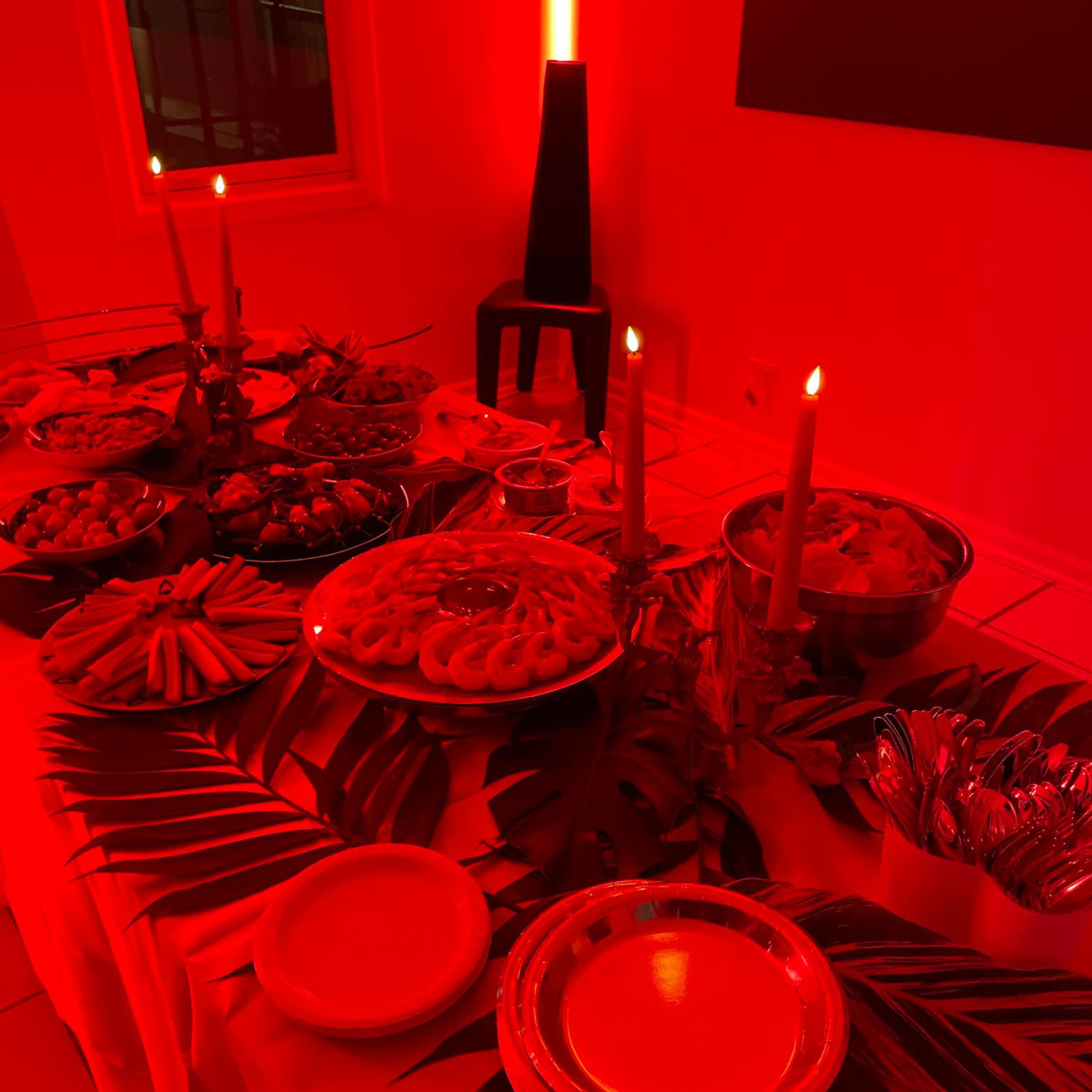 A table is set with various dishes and snacks, surrounded by red lighting and adorned with tropical leaves.
