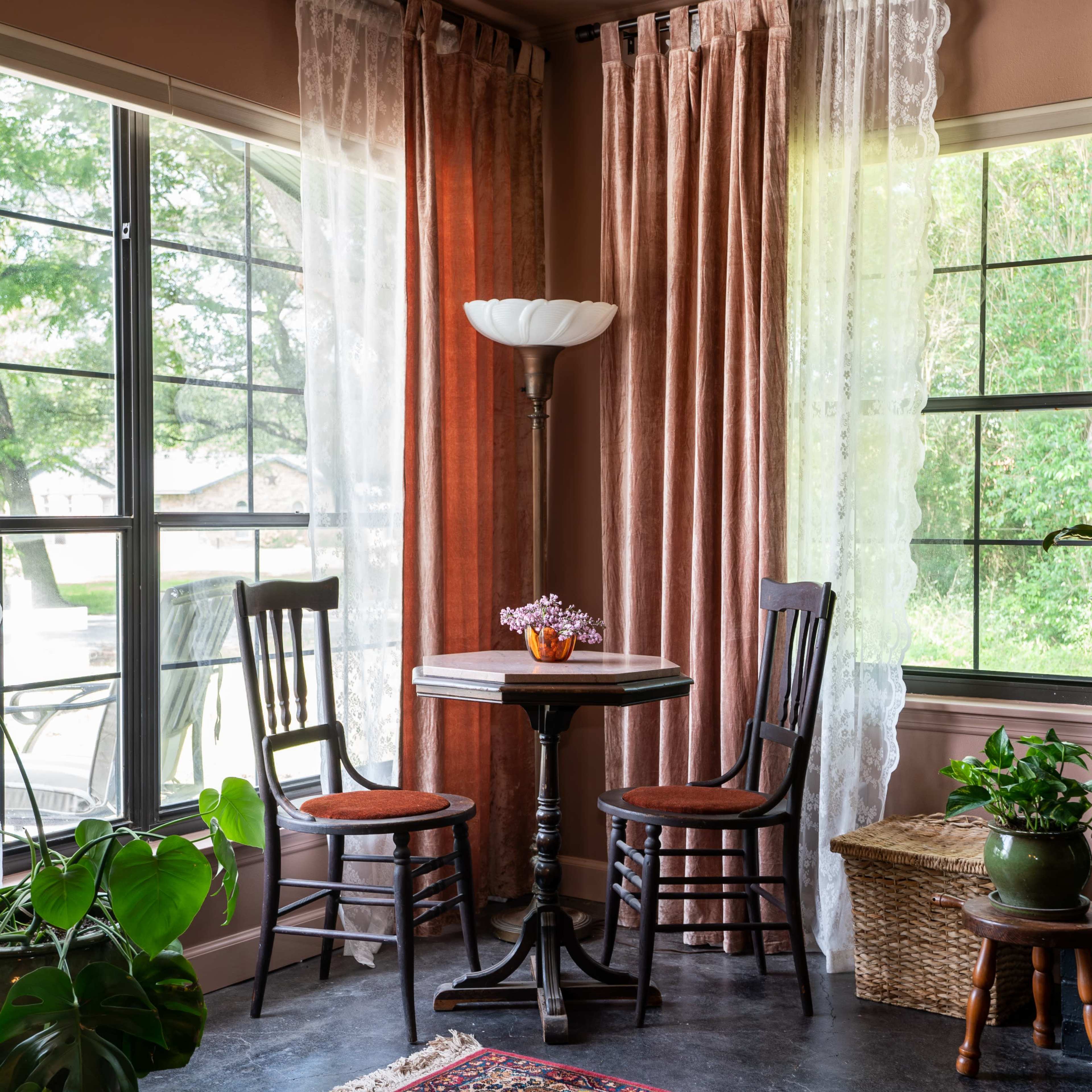 A small table with two chairs sits beside large windows draped with sheer and curtain panels, surrounded by indoor plants and a floral arrangement.