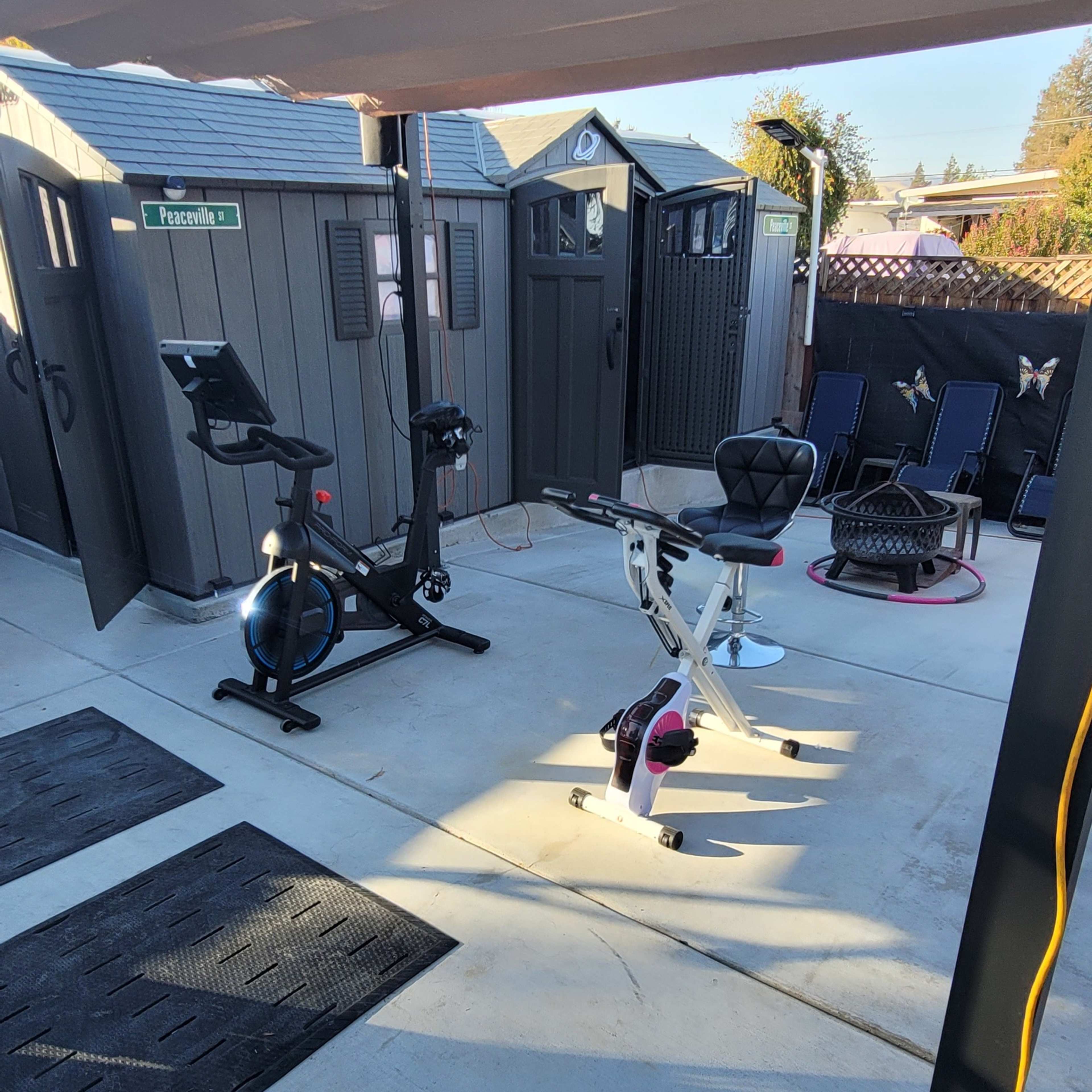 A home gym setup outside, featuring exercise equipment, including a stationary bike and a multi-use exercise bench, on a concrete floor.