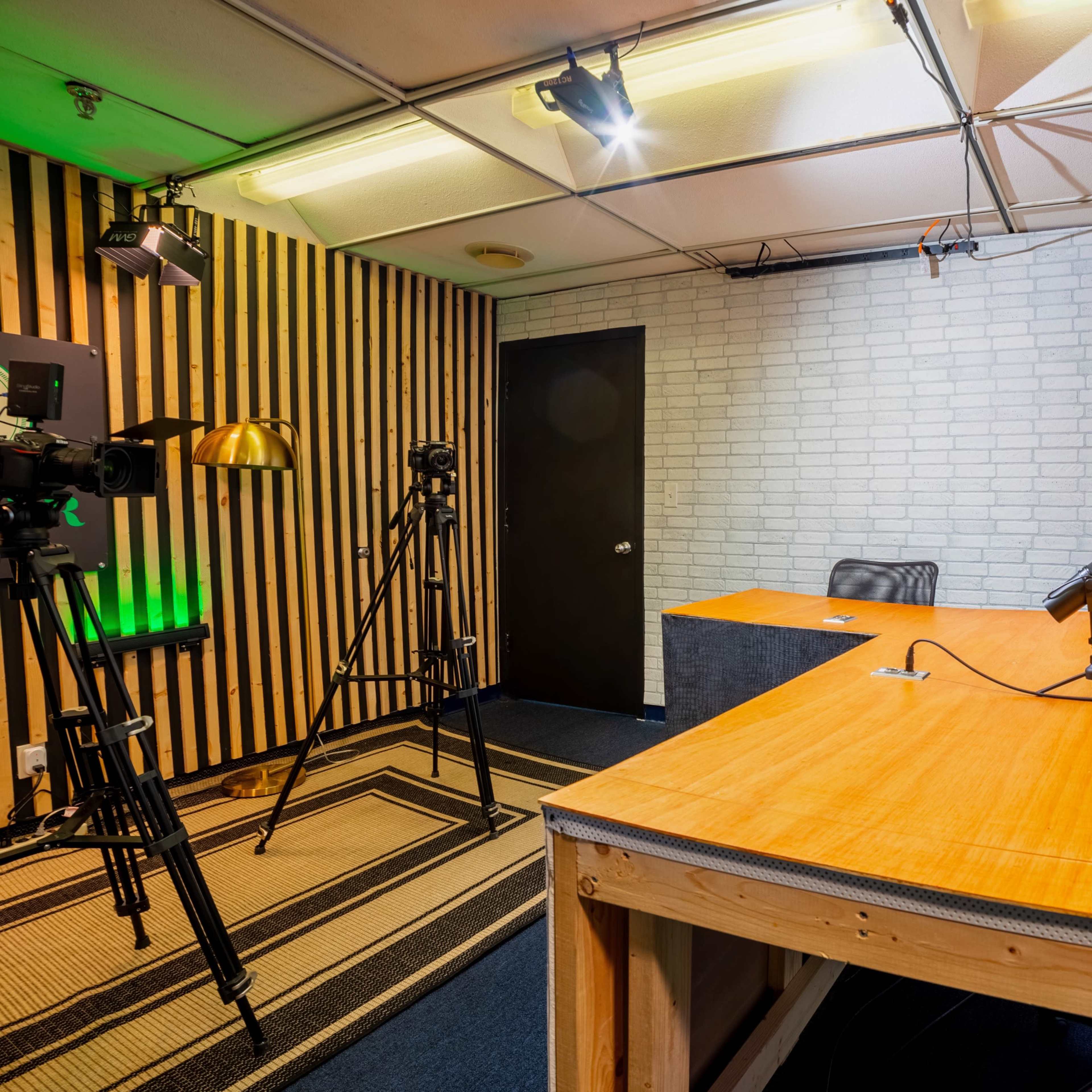 A video recording studio featuring a wooden desk, a microphone, camera equipment, and green plants against a textured wall.