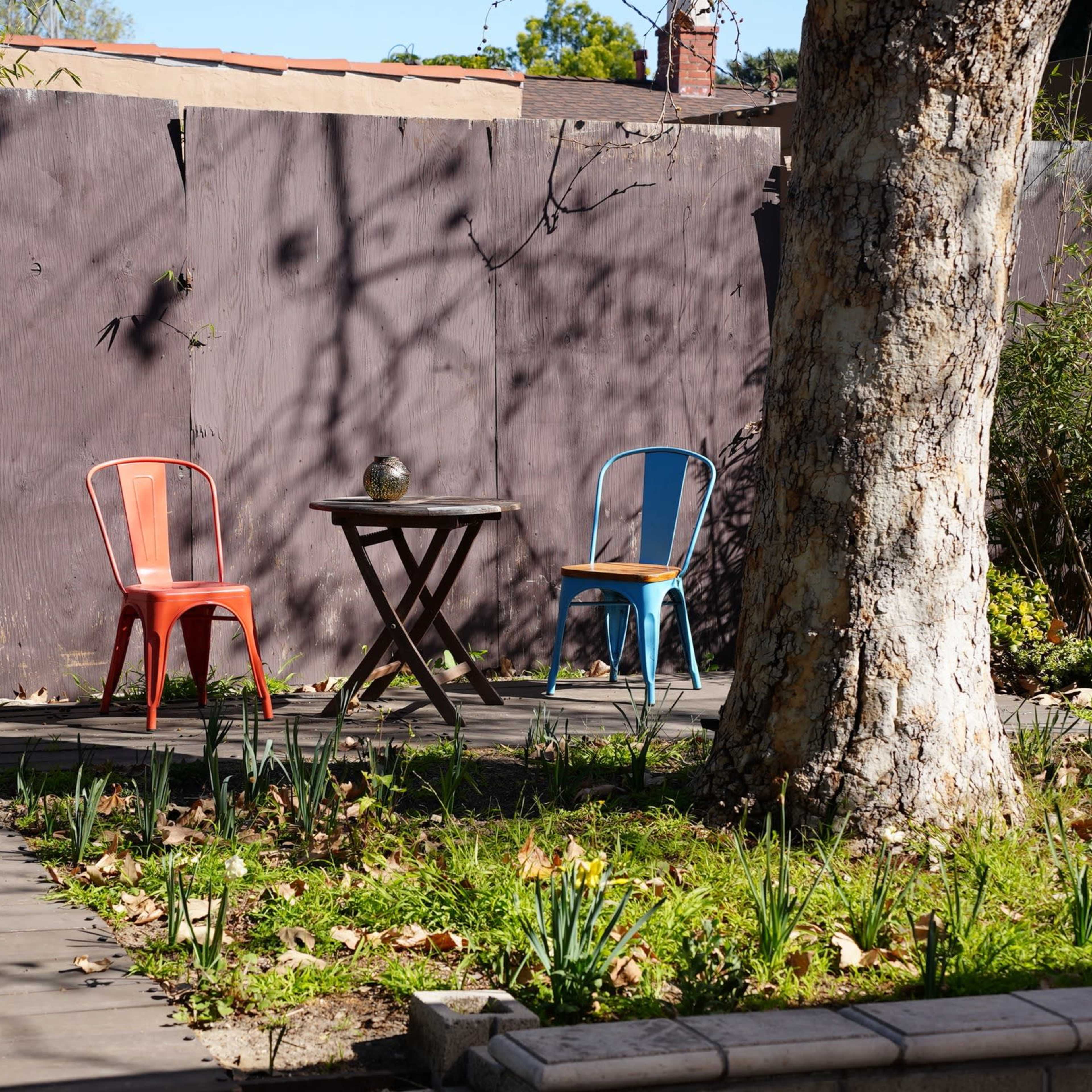A small outdoor patio features a wooden table surrounded by two chairs, one red and one blue, with a backdrop of a purple wall and greenery.