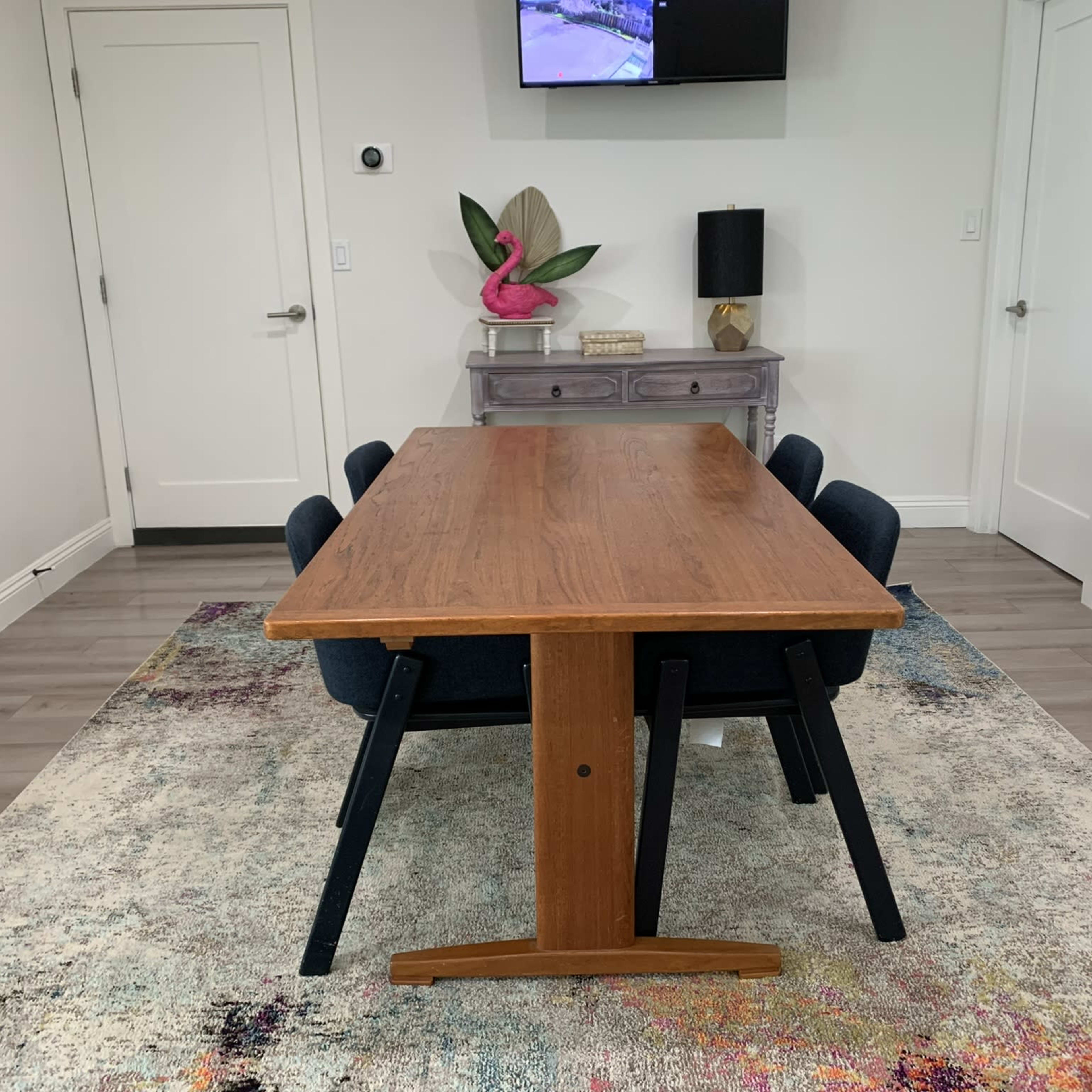 A wooden dining table is set in a room with blue chairs, a colorful rug, and a television mounted on the wall.
