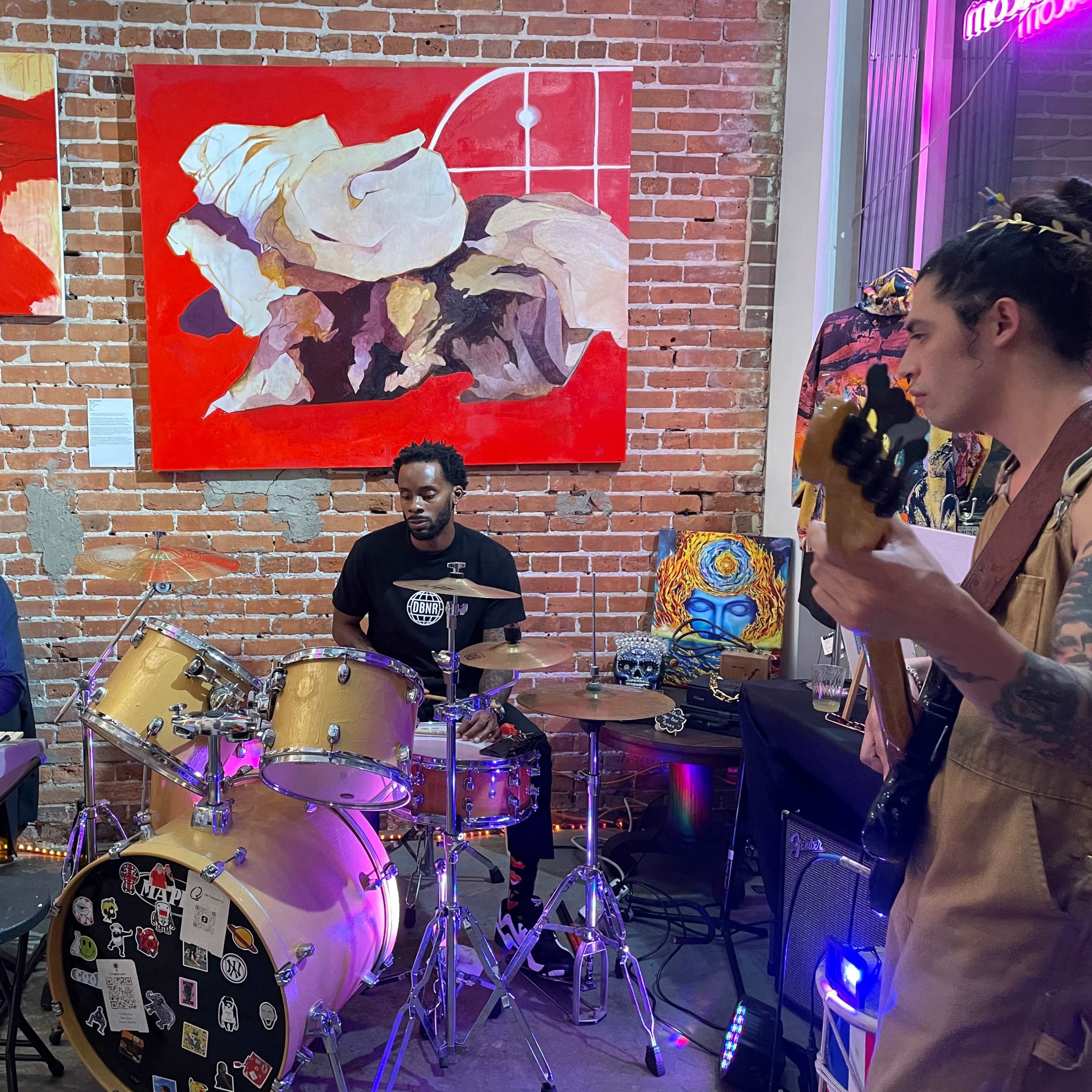 A trio of musicians performs in a brick-walled venue, featuring a keyboardist, a drummer, and a bassist, with colorful artwork displayed in the background.