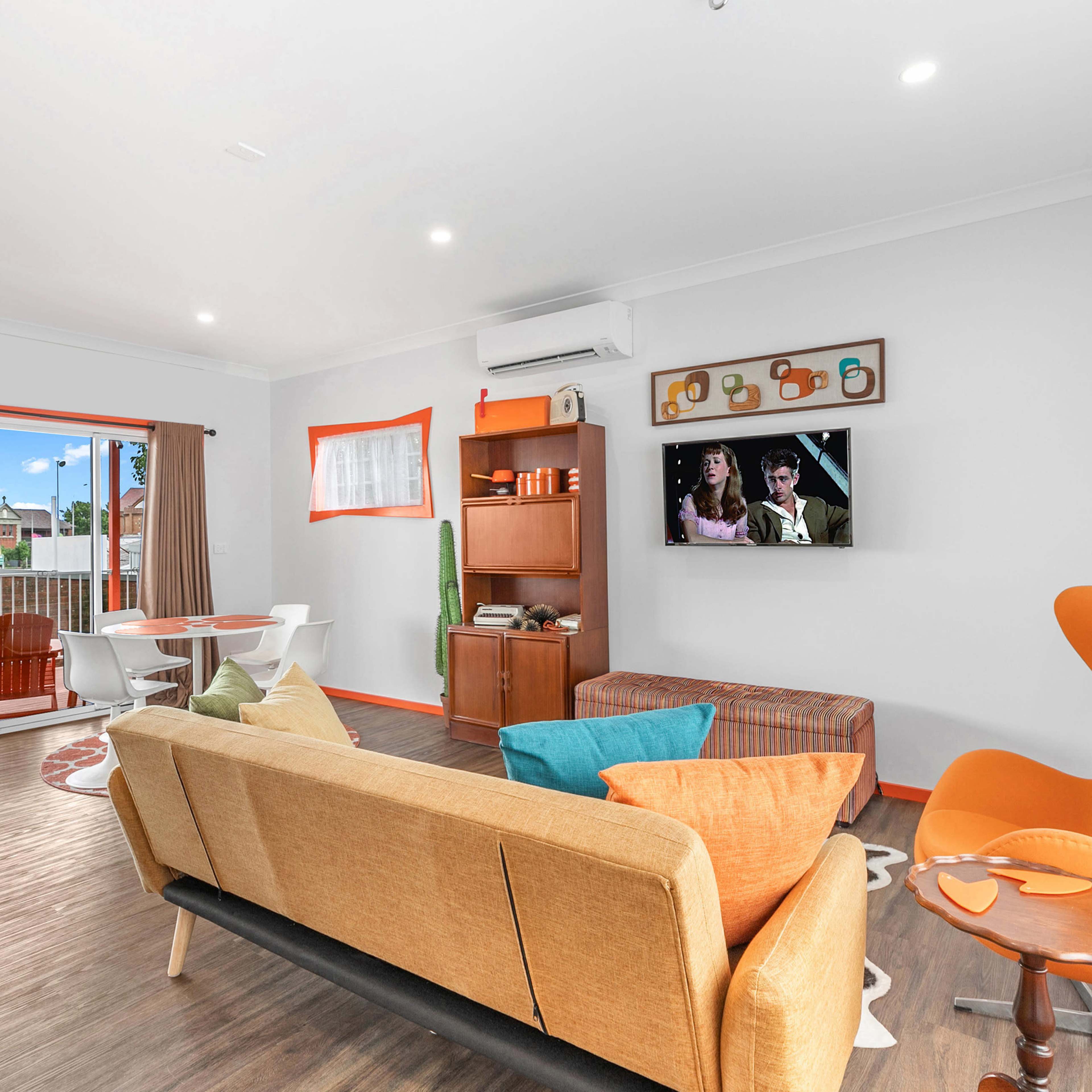 A modern living room features a sofa, an orange armchair, a television on the wall, and a dining area with a round table and chairs.