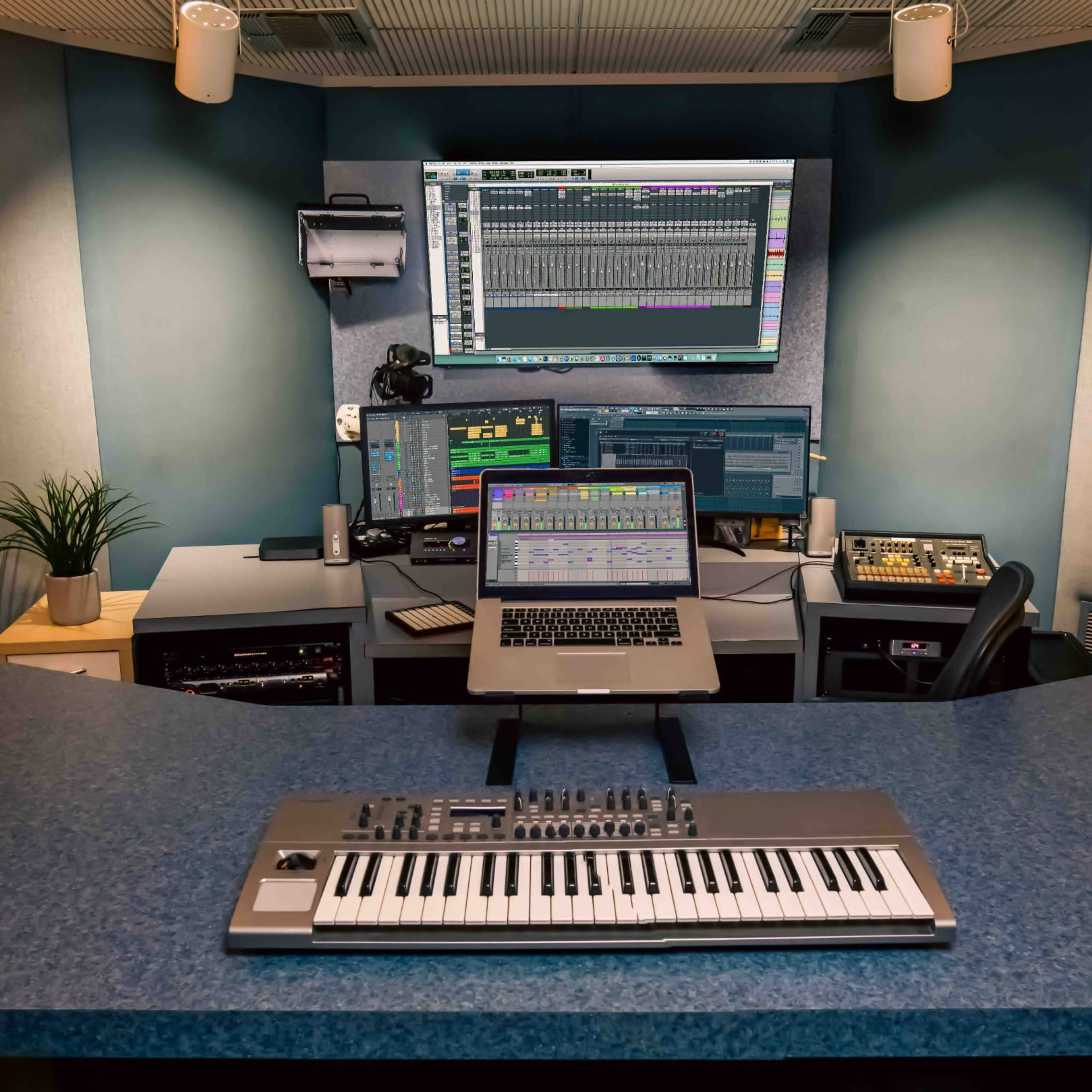 A music production studio with a computer setup, multiple monitors, a keyboard, and a plant on a desk.