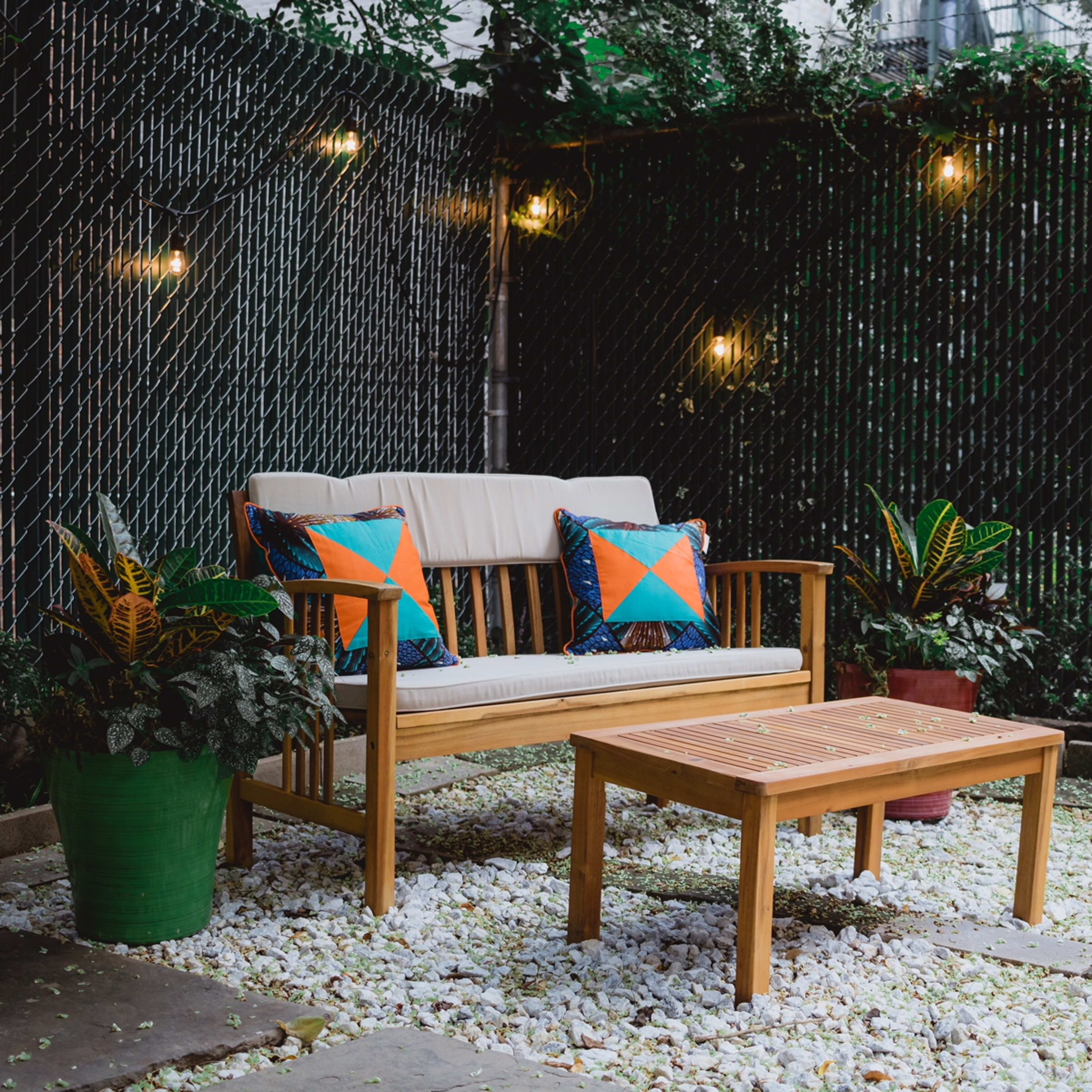 A wooden sofa with colorful cushions and a matching coffee table sits in a gravel-covered patio surrounded by potted plants and a chain-link fence.