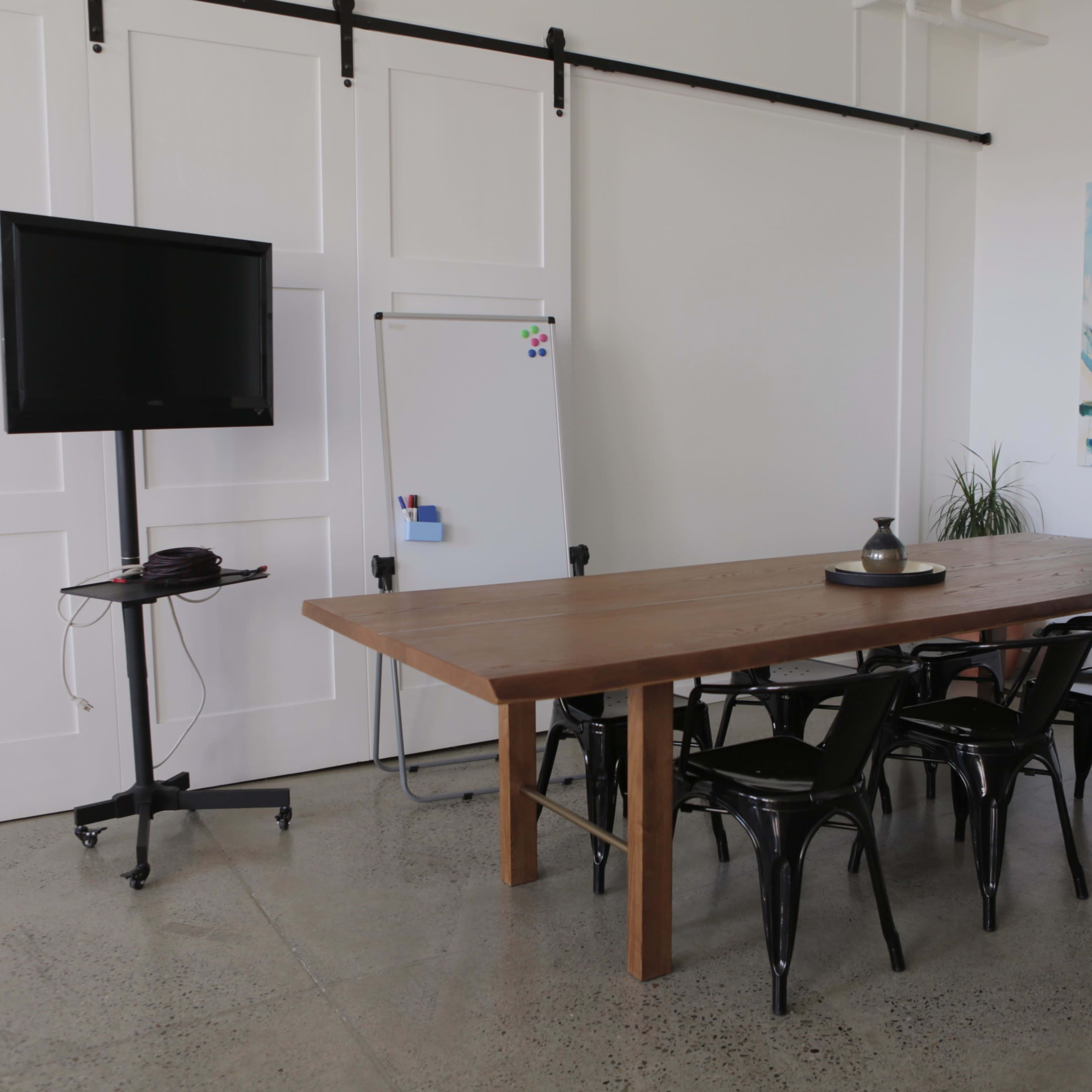 A conference room features a wooden table surrounded by black chairs, a television on a stand, a whiteboard, and a piece of wall art.