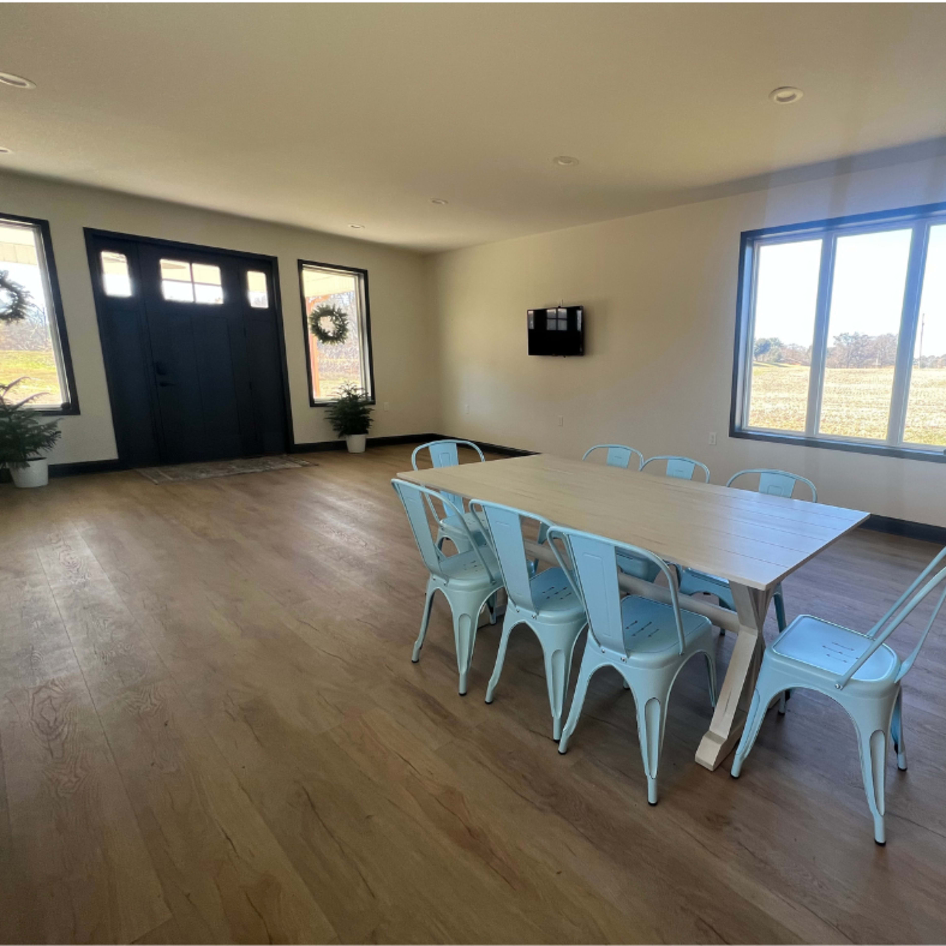 A spacious room features a wooden dining table surrounded by metal chairs, with large windows and a front door visible in the background.