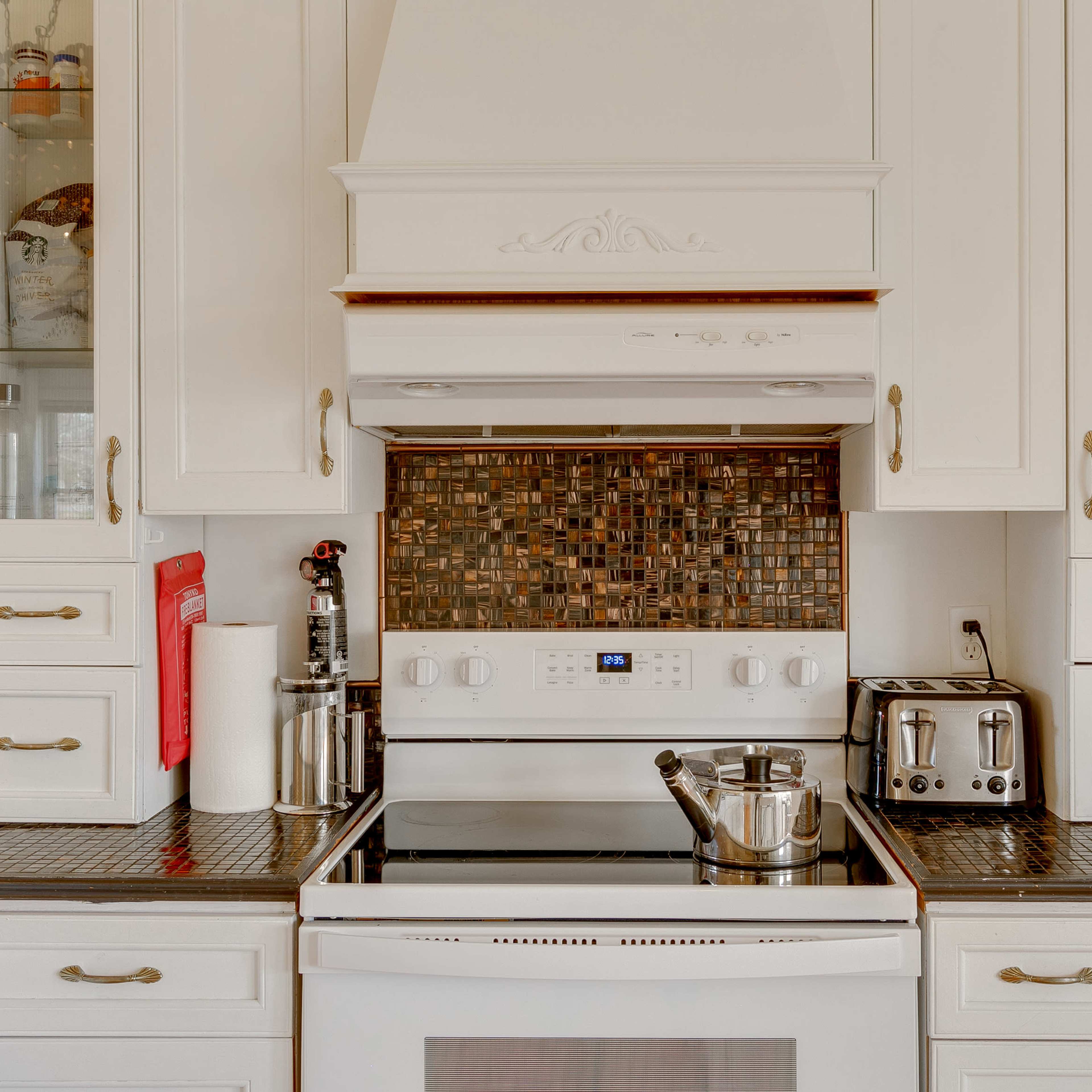 A modern kitchen features a stove beneath a range hood, with a tiled backsplash, coffee maker, toaster, and cabinets.