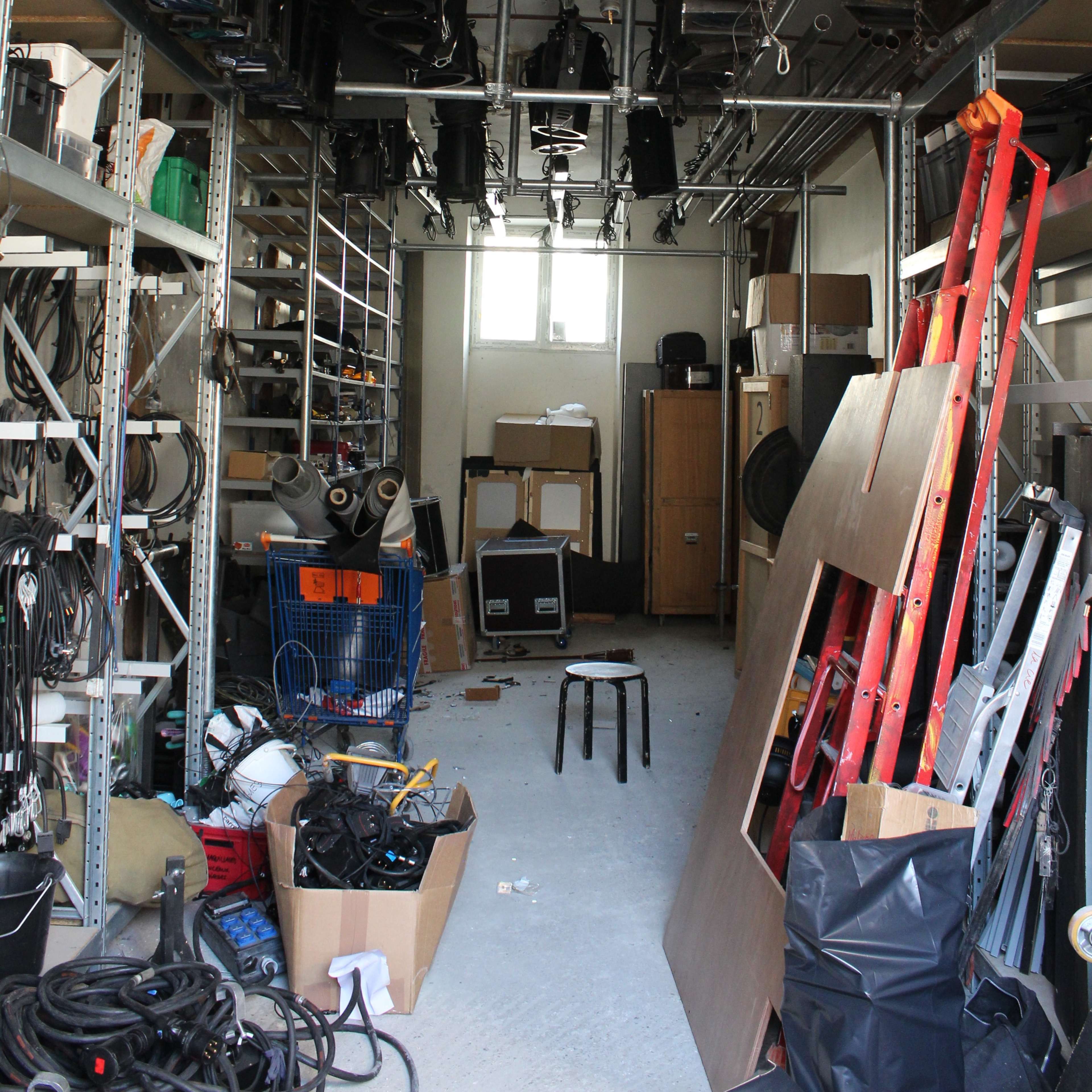 A storage room filled with hanging lighting equipment, shelves of tools, and scattered cables and materials.