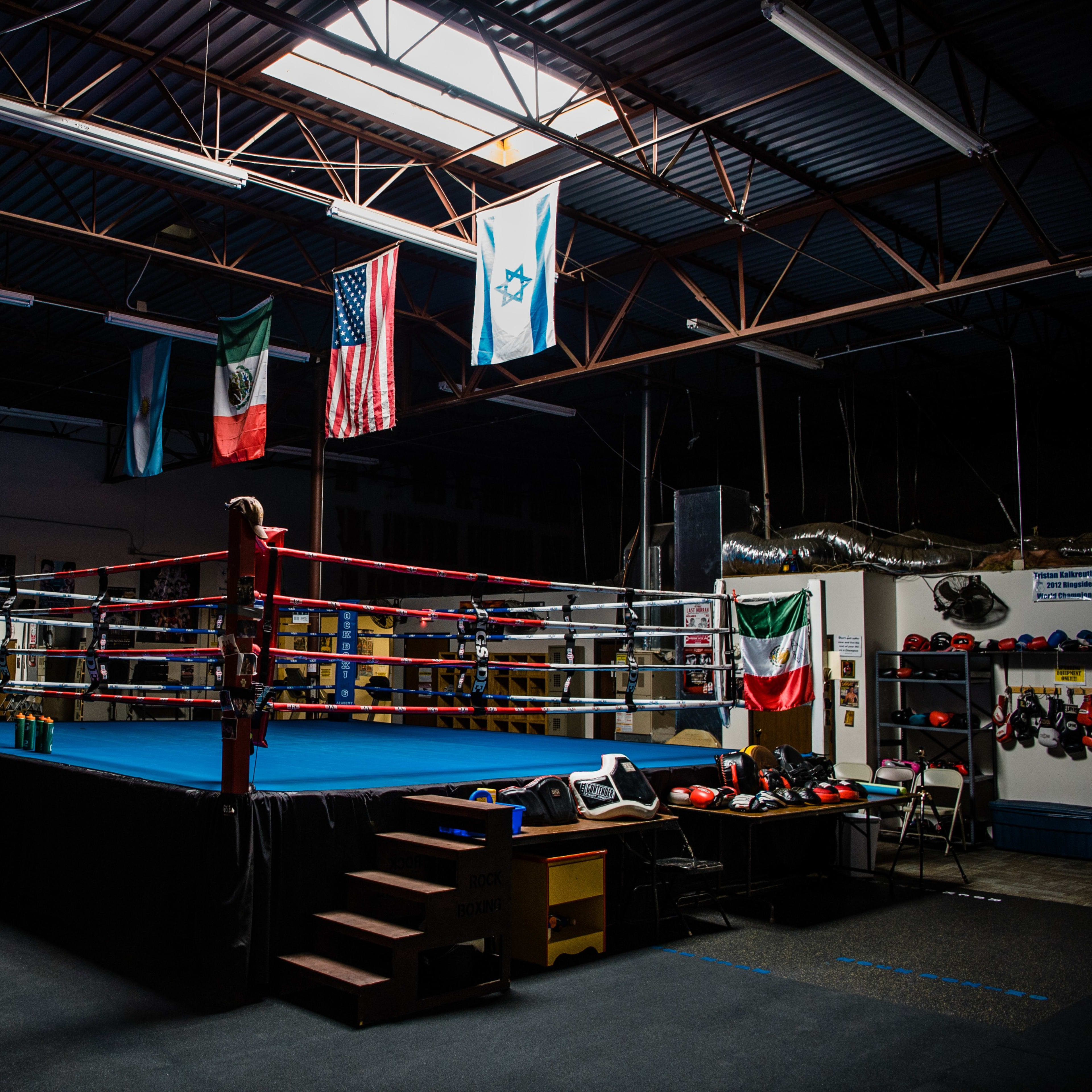 A boxing gym with a blue-ringed boxing ring, several flags hanging from the ceiling, and training equipment along the walls.