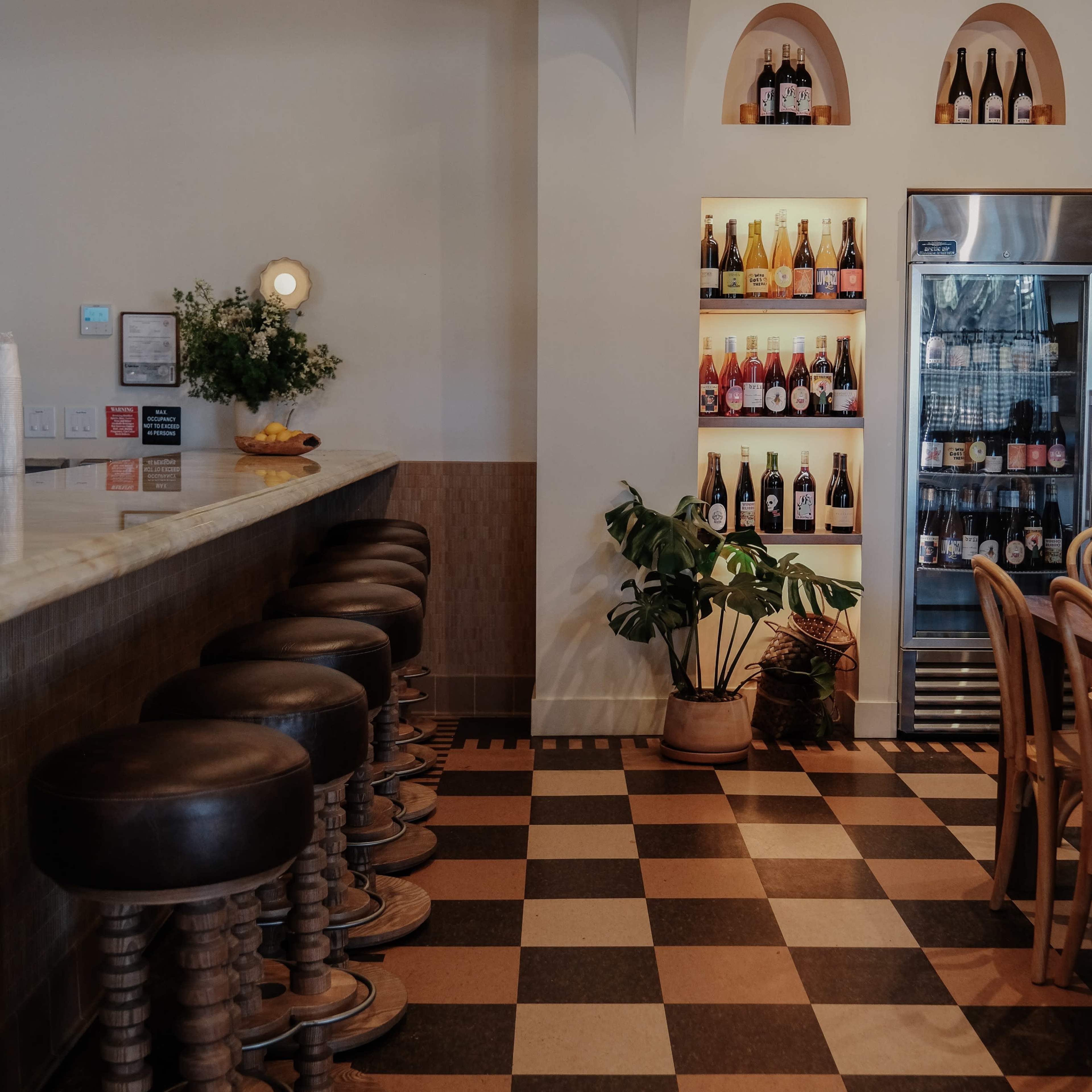 A bar area with a long countertop, wooden stools, a plant, and a wall displaying various bottles in a refrigerator and on shelves.