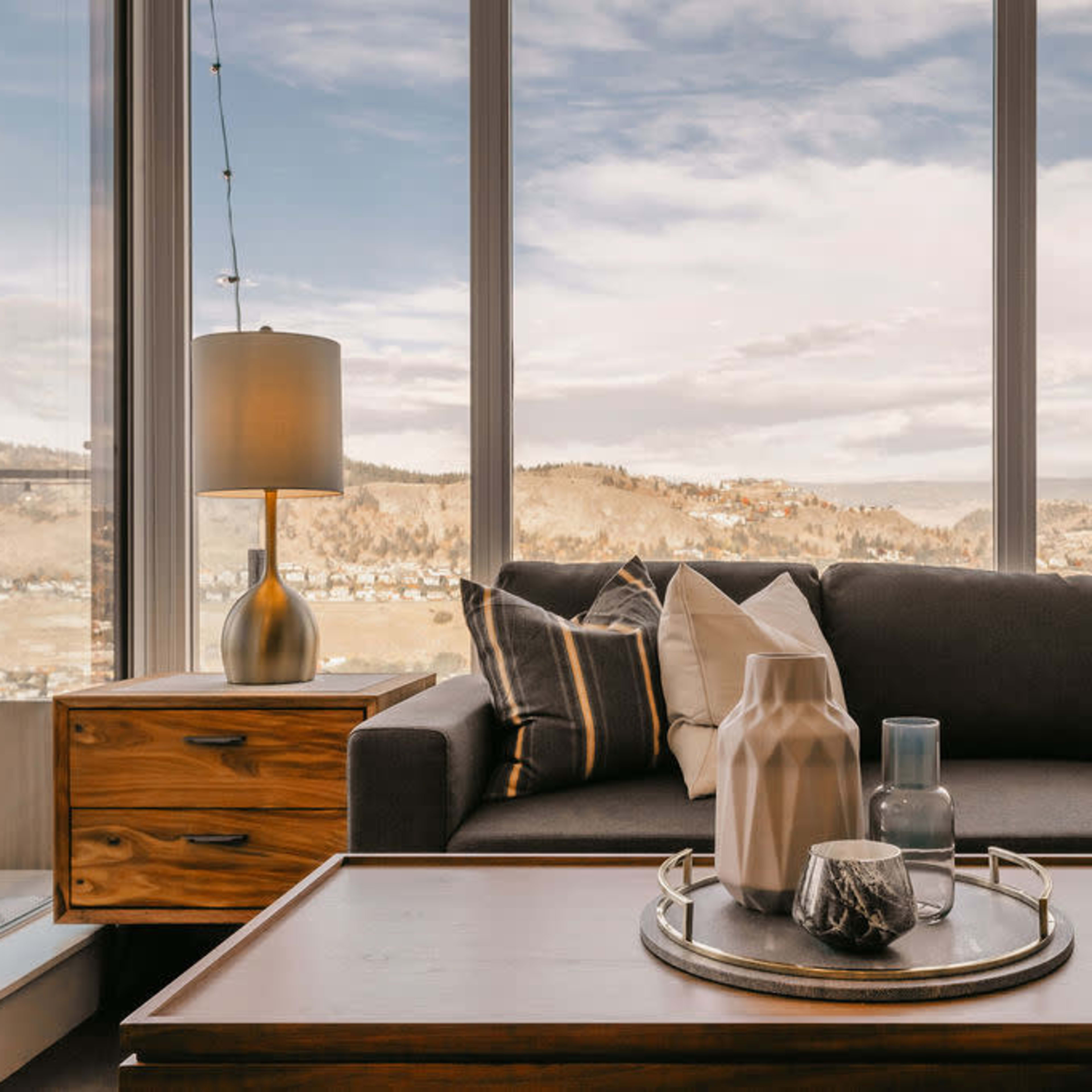 The interior features a modern living room with a gray sofa, a wooden side table, and large windows showing a view of mountains and a cloudy sky.