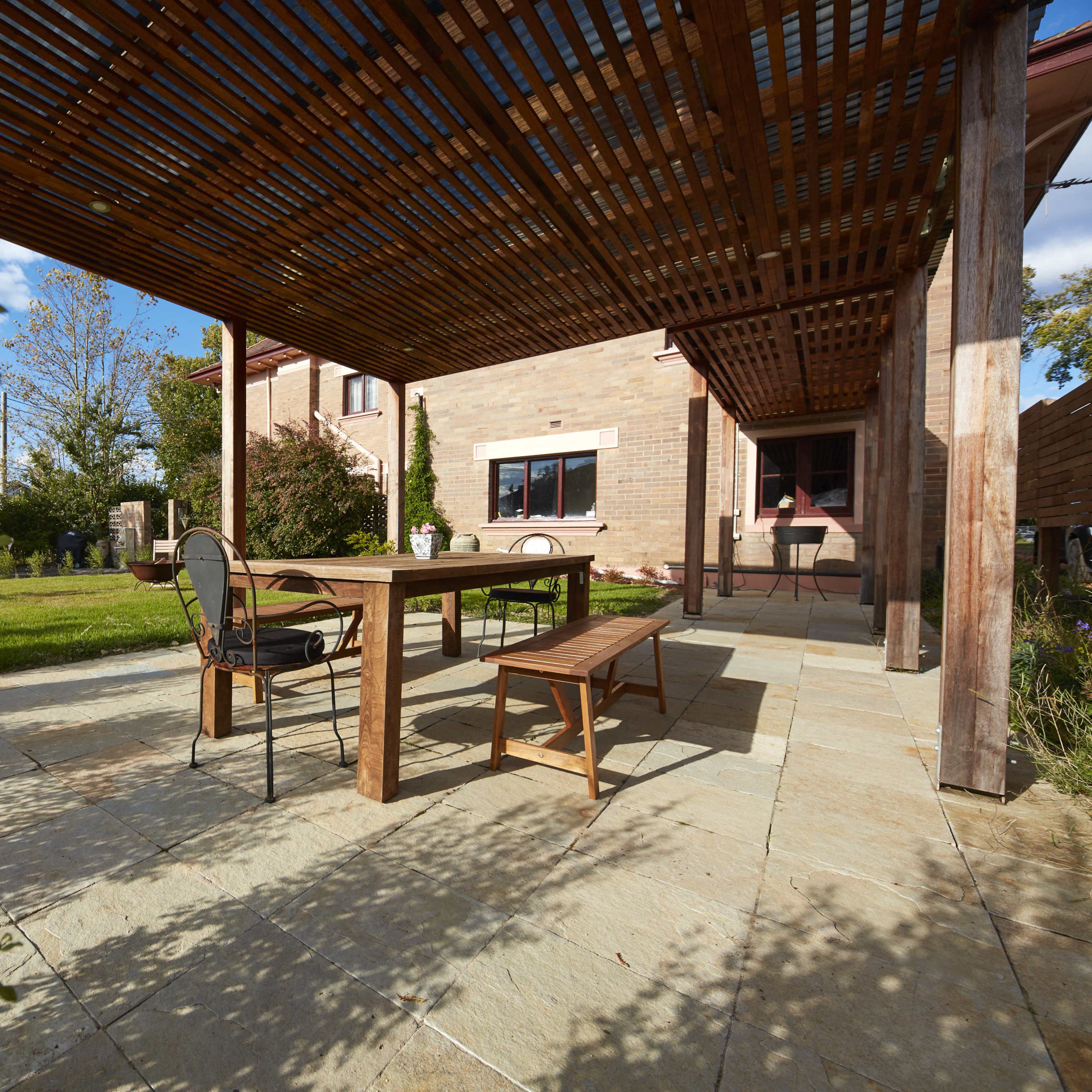 A wooden table and bench are set under a pergola on a stone patio beside a house surrounded by greenery.