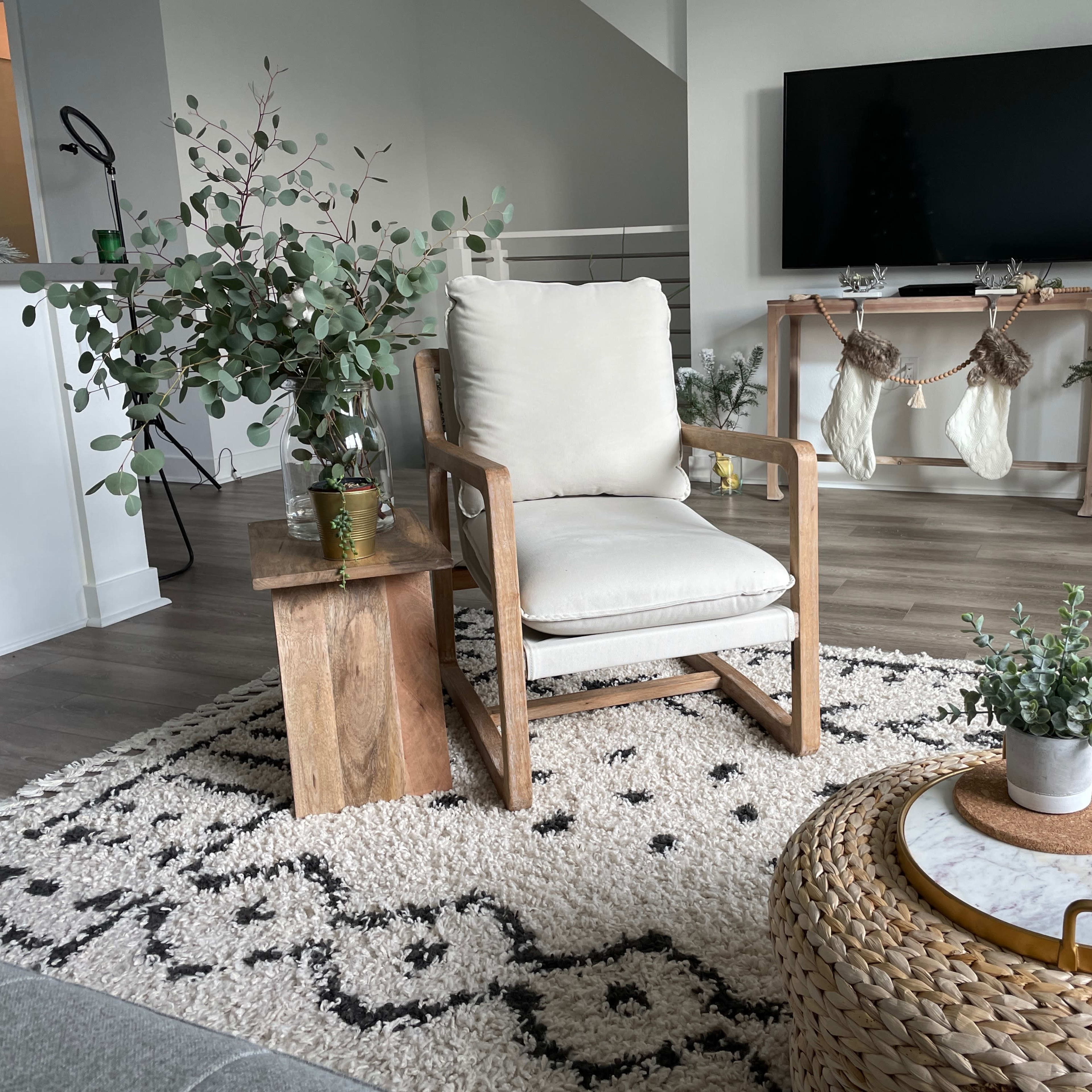 The scene features a cozy living room with a wooden chair, a small side table, a round coffee table with a marble top, and festive decorations along with plants on a textured rug.
