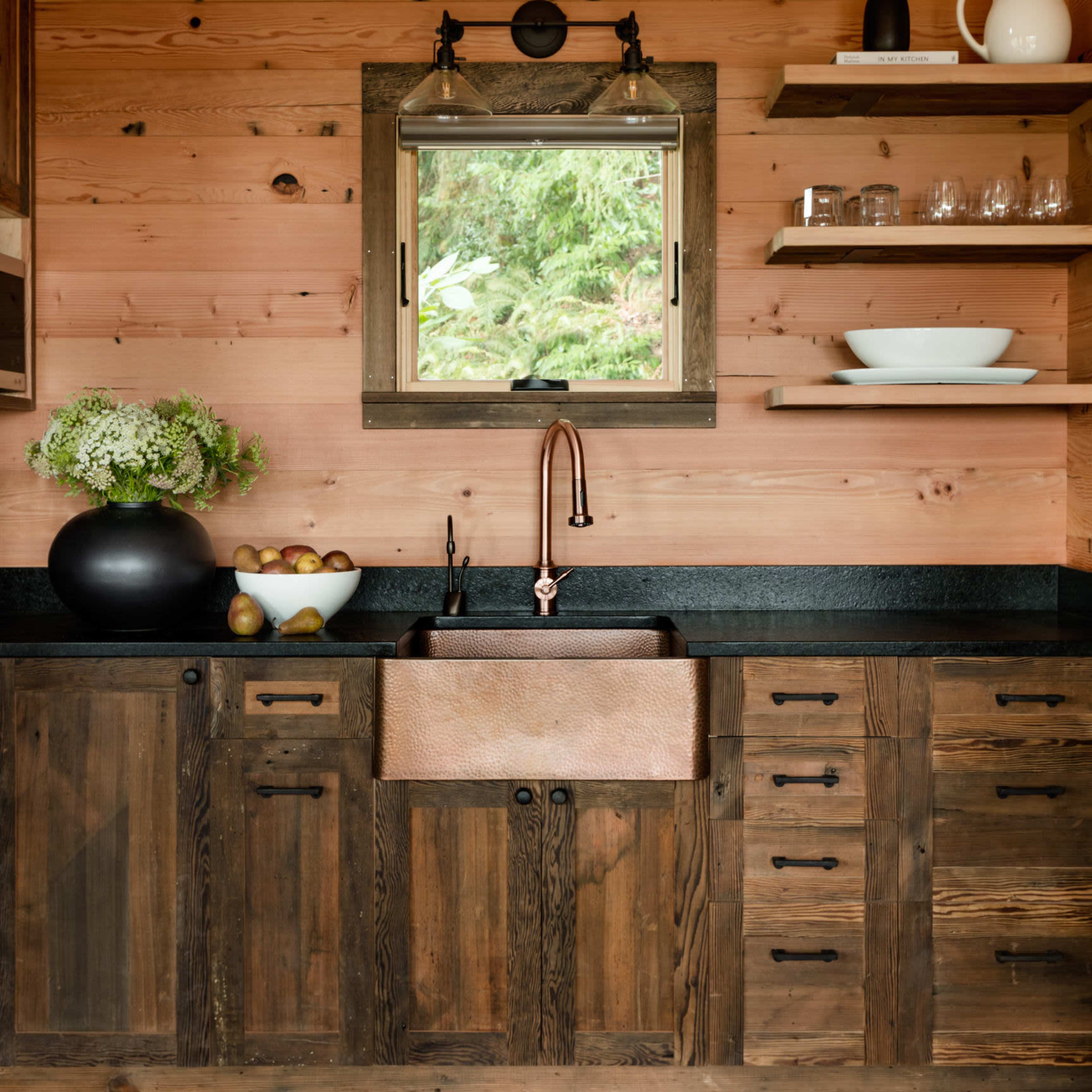 A rustic kitchen with a large copper farmhouse sink beneath a window, wooden cabinetry, and open shelving displaying dishes and decorative items.