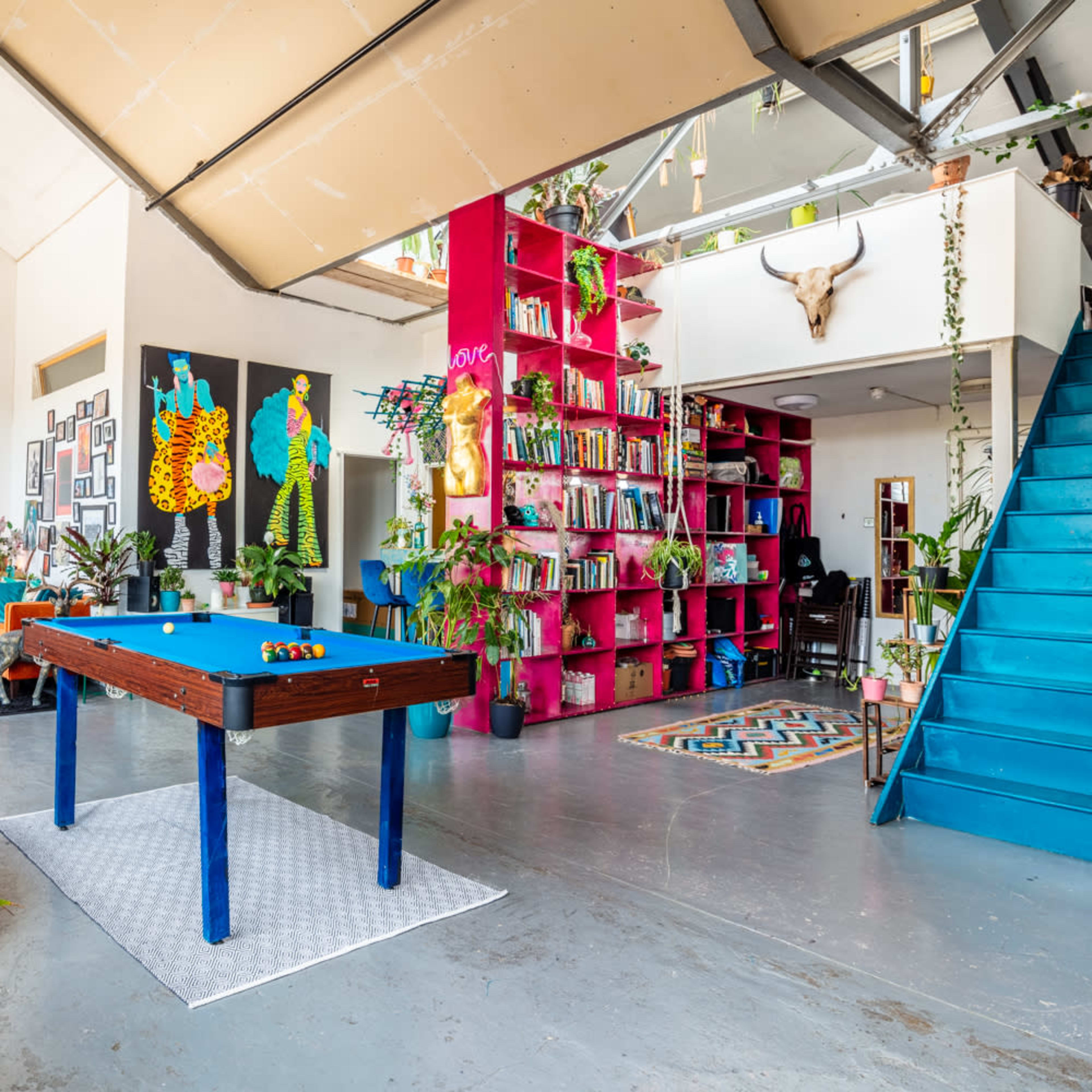 A vibrant interior space with a blue pool table in the center, surrounded by colorful decor, bookshelves, and a staircase leading to an upper level.