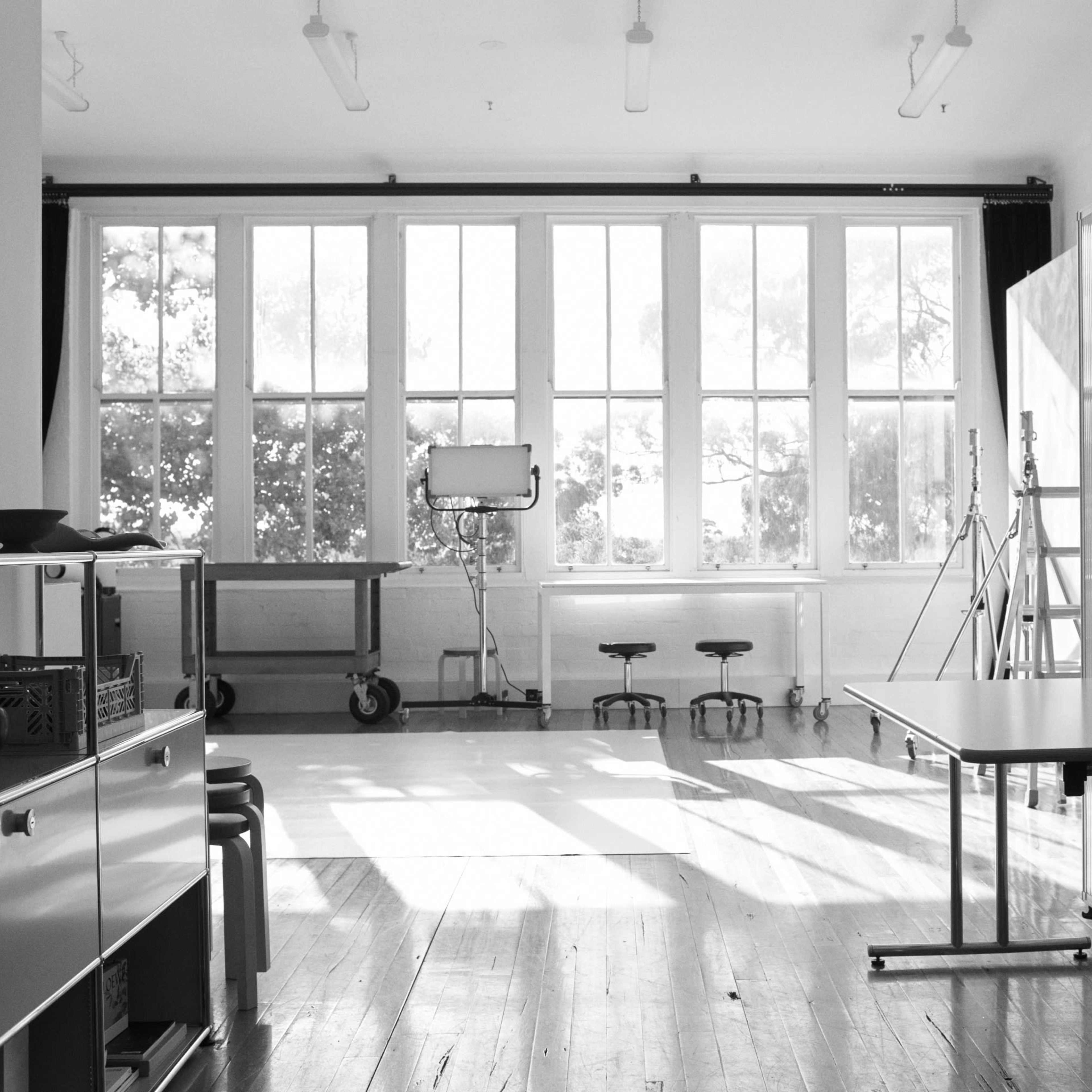 A spacious, empty room with large windows, light wood flooring, and a few pieces of furniture including tables and stools.