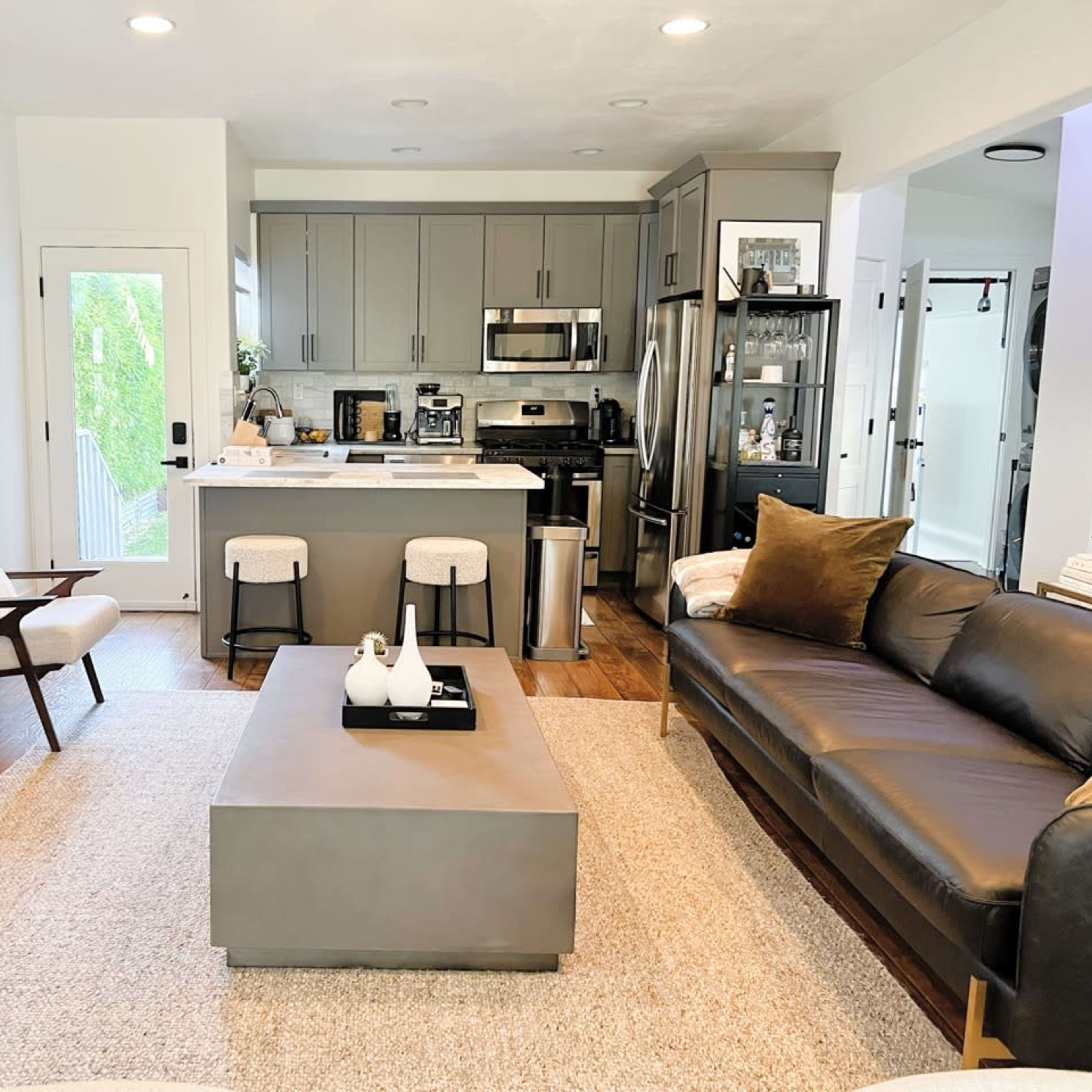 A modern living room with a sofa, armchair, coffee table, and a kitchen area in the background, featuring stainless steel appliances and cabinetry.