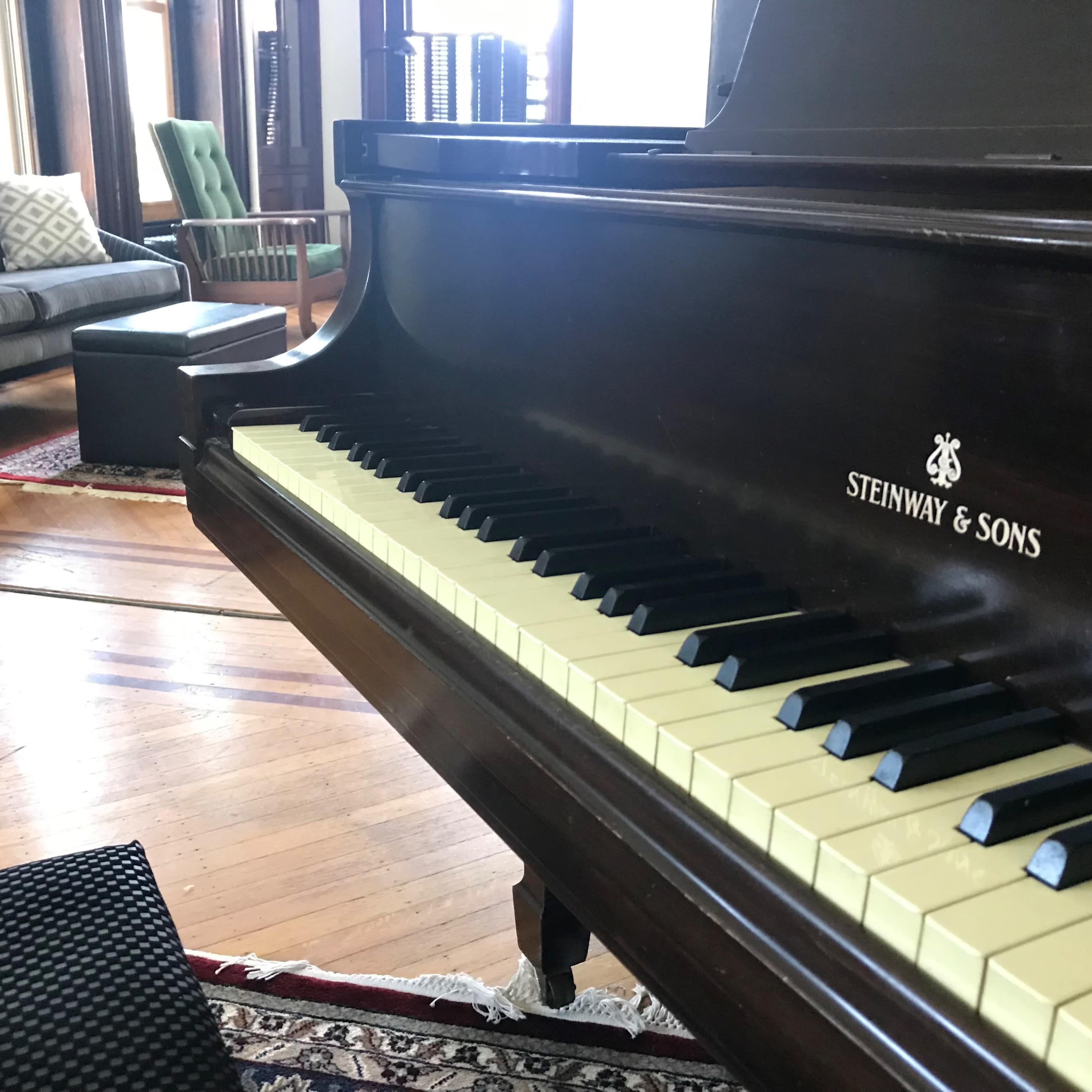 A Steinway & Sons piano is positioned in a room featuring a couch and a patterned rug on wooden floors.