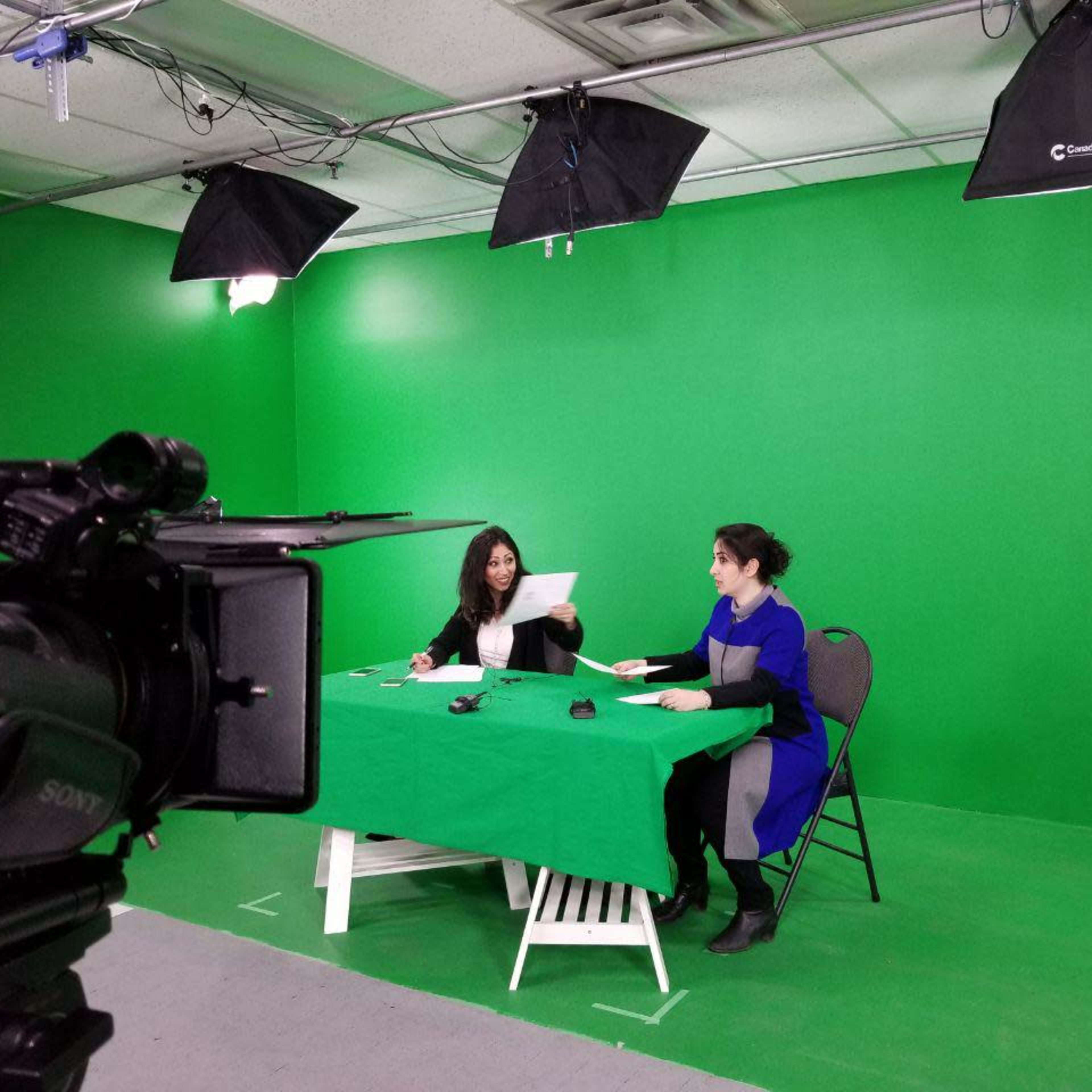 Two women sit at a table covered with a green cloth in front of a green screen, preparing for a video shoot under multiple studio lights.