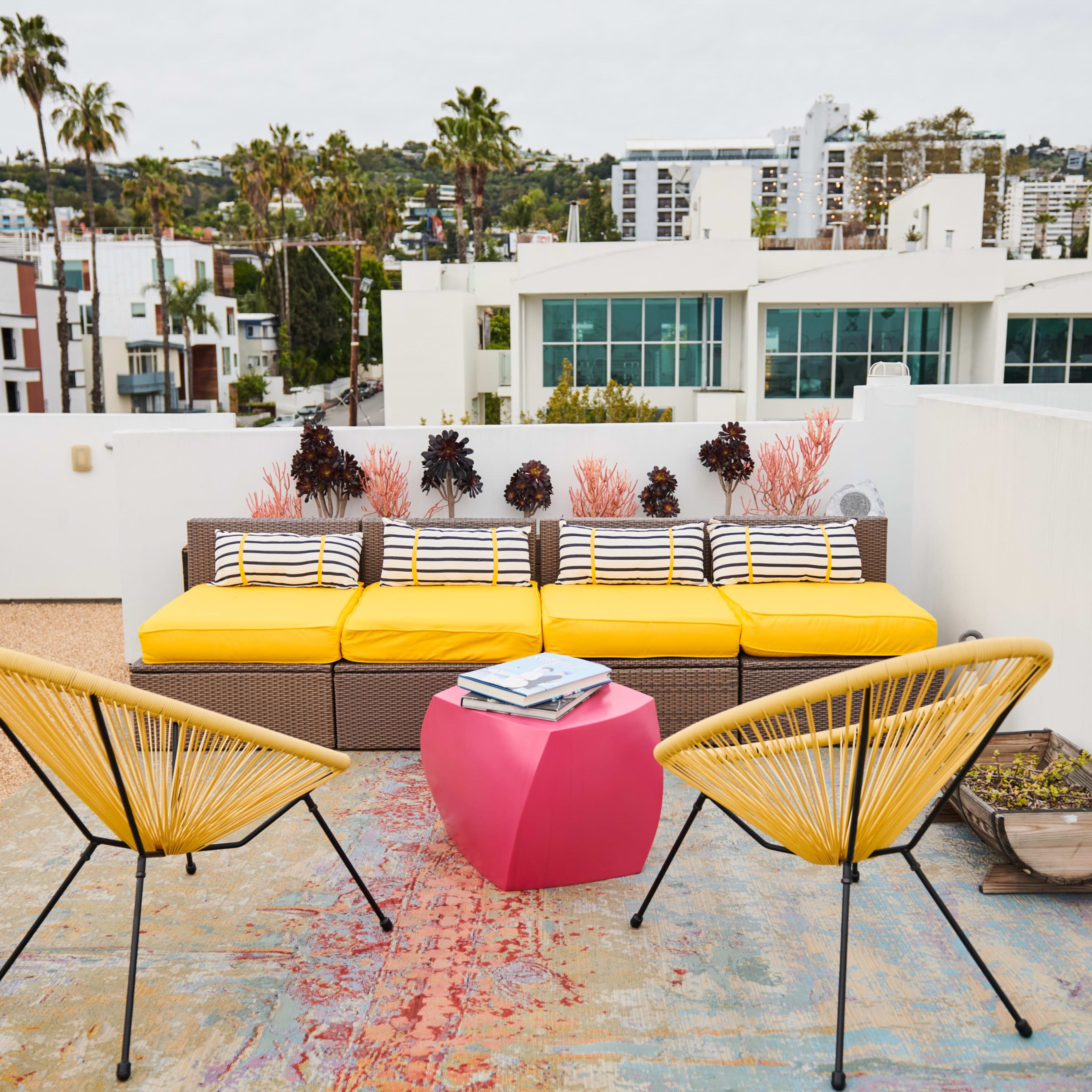 A rooftop terrace featuring a yellow couch with striped cushions, two black chairs, a pink side table, and potted plants, against a backdrop of buildings and palm trees.