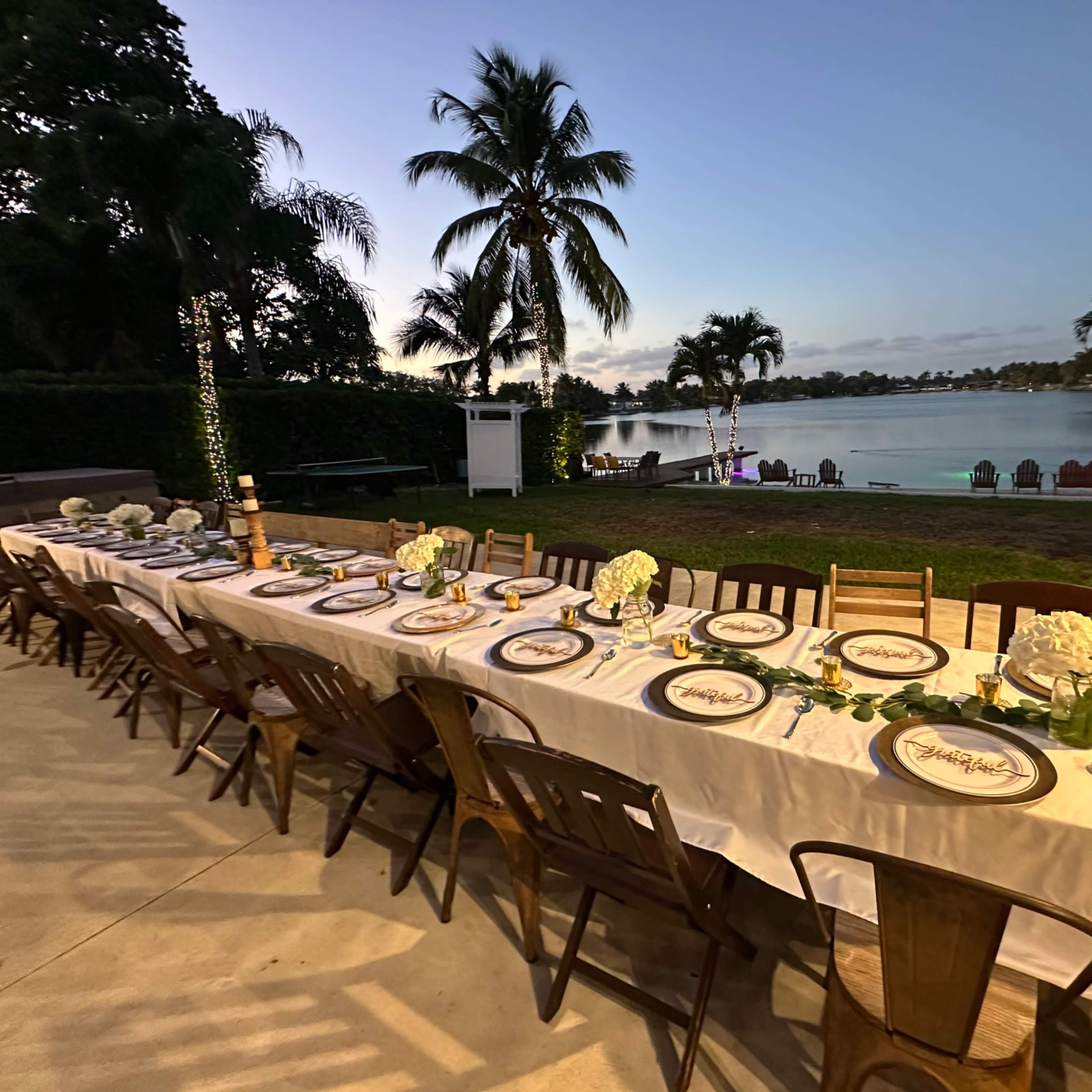 A long dining table is set for a gathering beside a lake, flanked by palm trees and a patio area.