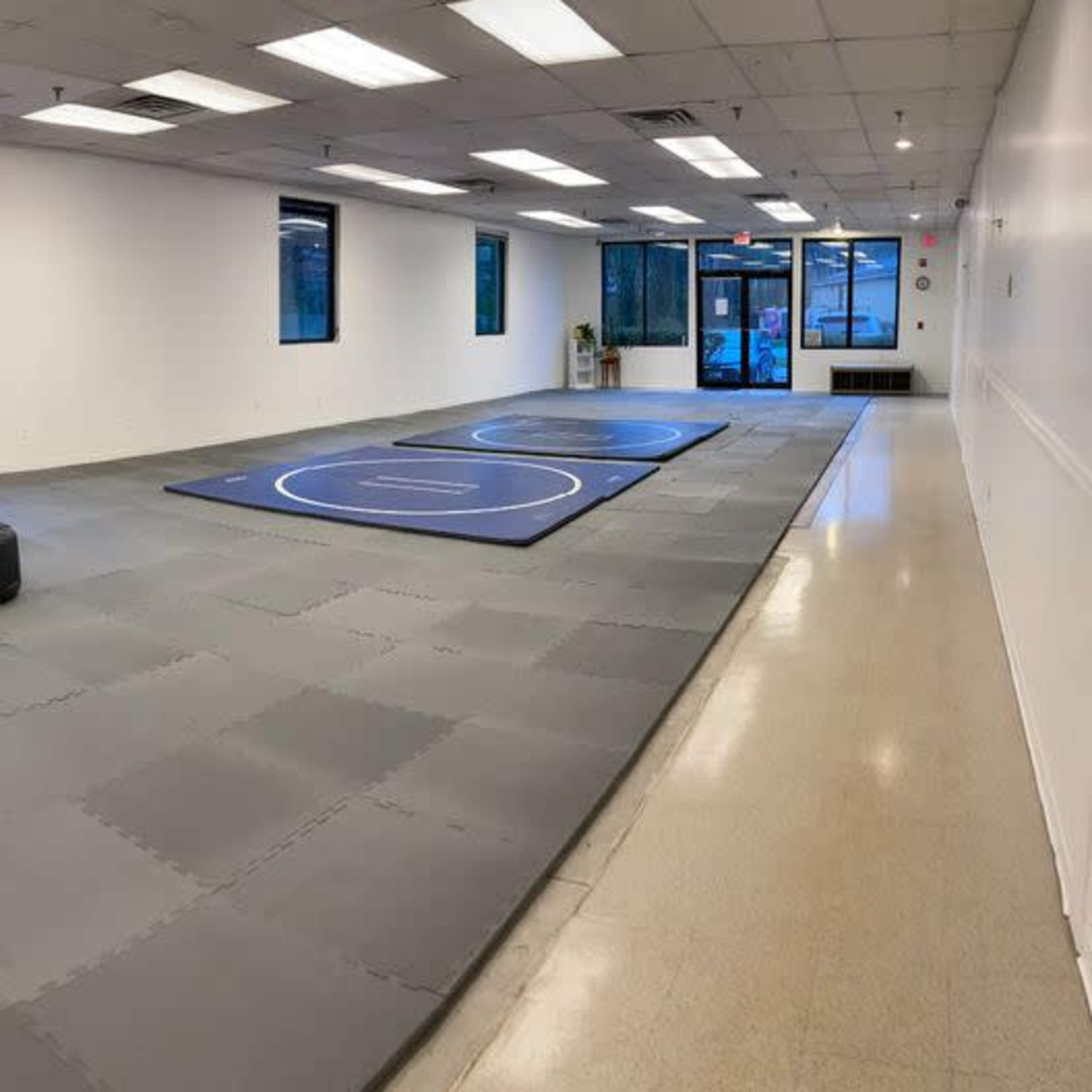 A spacious, empty training room with gray mats, two circular wrestling rings, and black practice equipment arranged on one side.