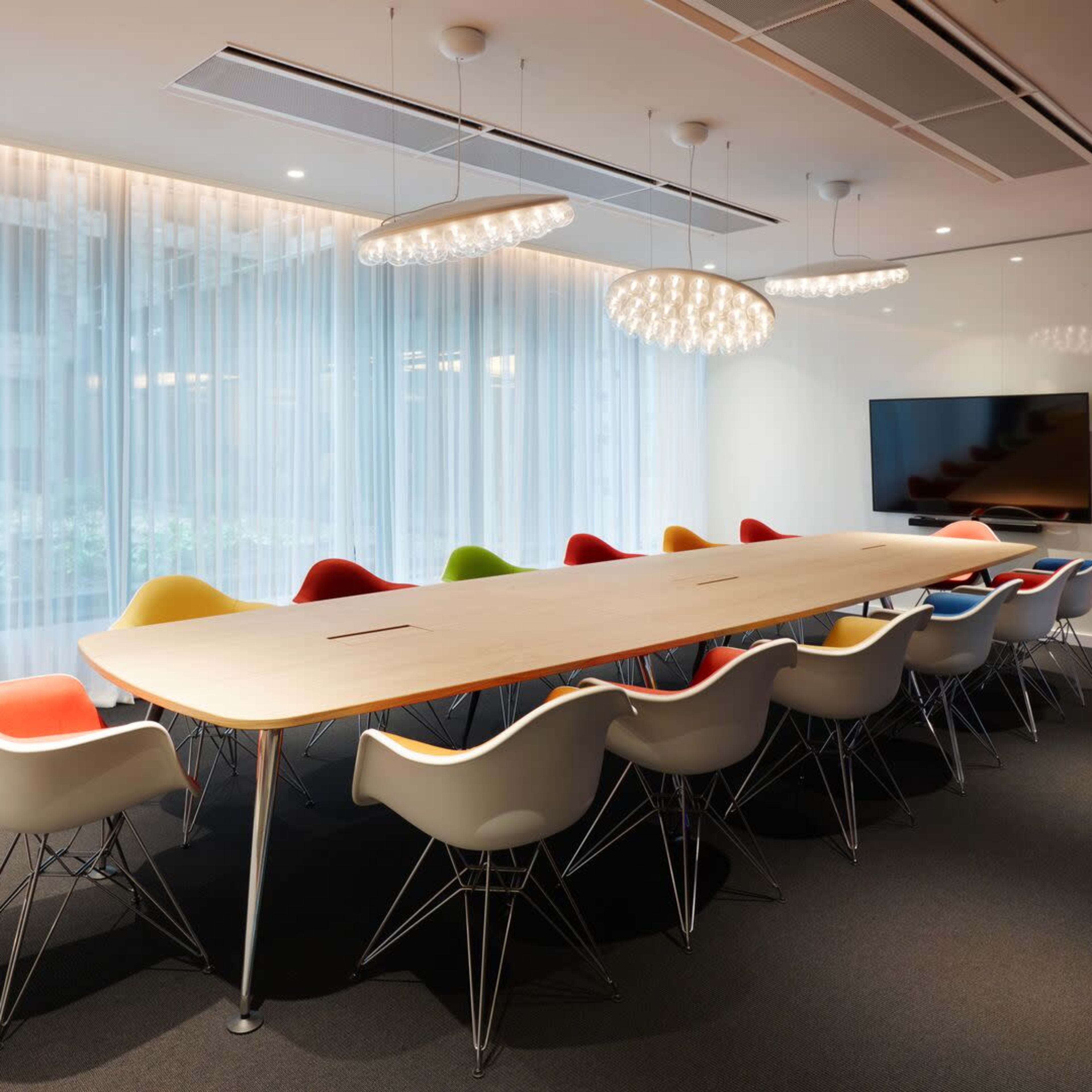A large conference room features a long wooden table surrounded by colorful Eames-style chairs and a wall-mounted television.
