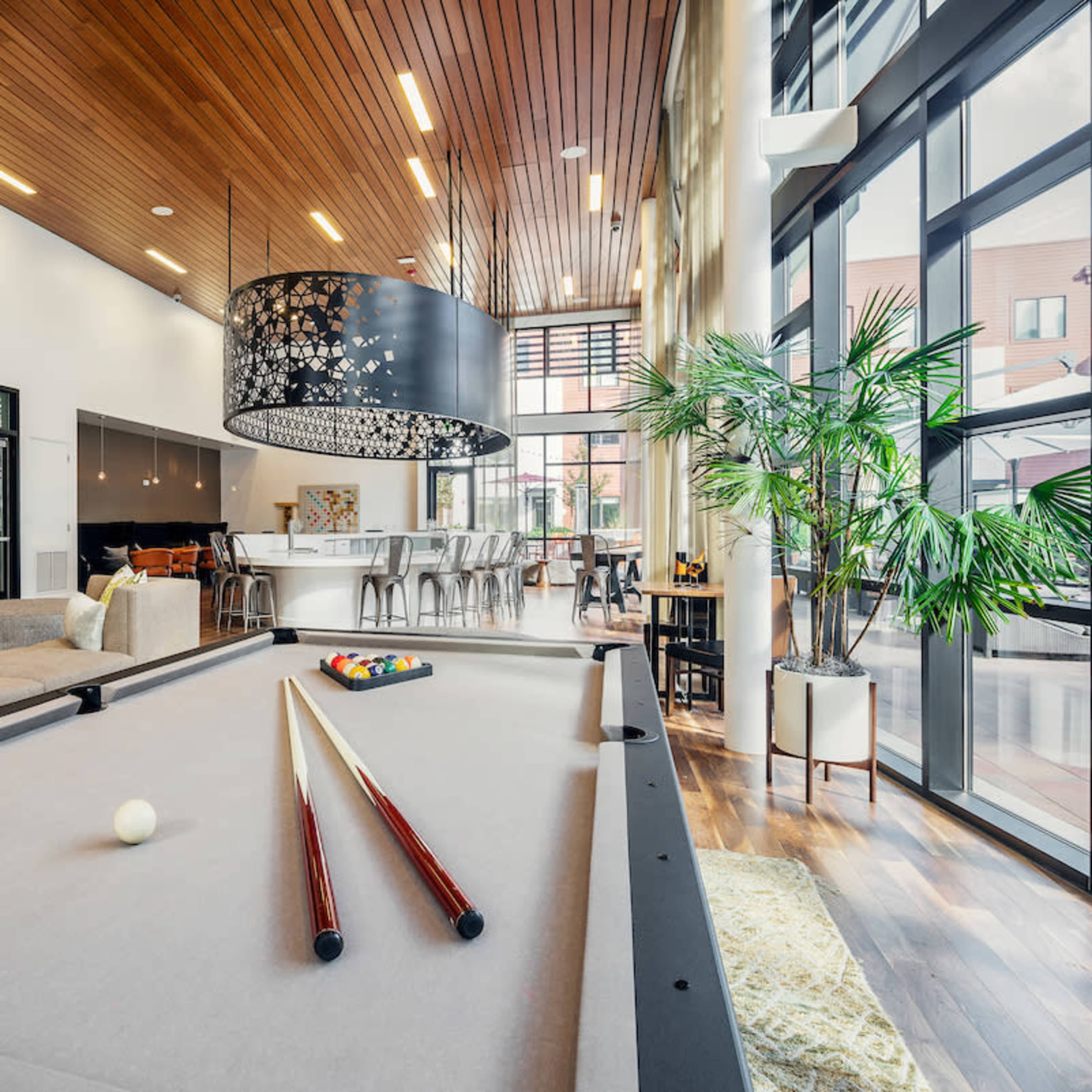 A spacious room features a pool table in the foreground, surrounded by contemporary furniture and large windows that allow ample natural light.