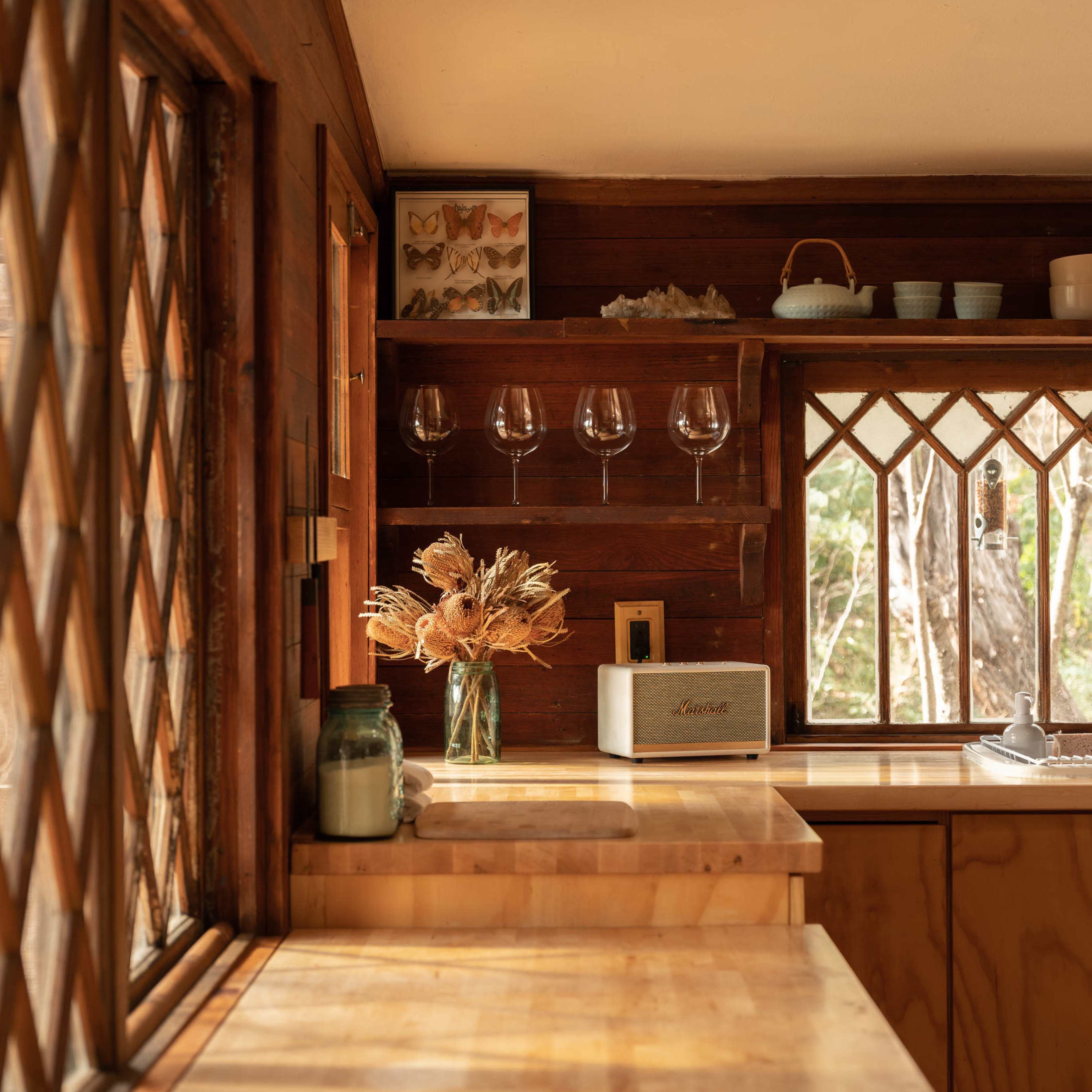 A light-filled kitchen features wooden countertops, a vase of dried flowers, and glassware displayed on a shelf.